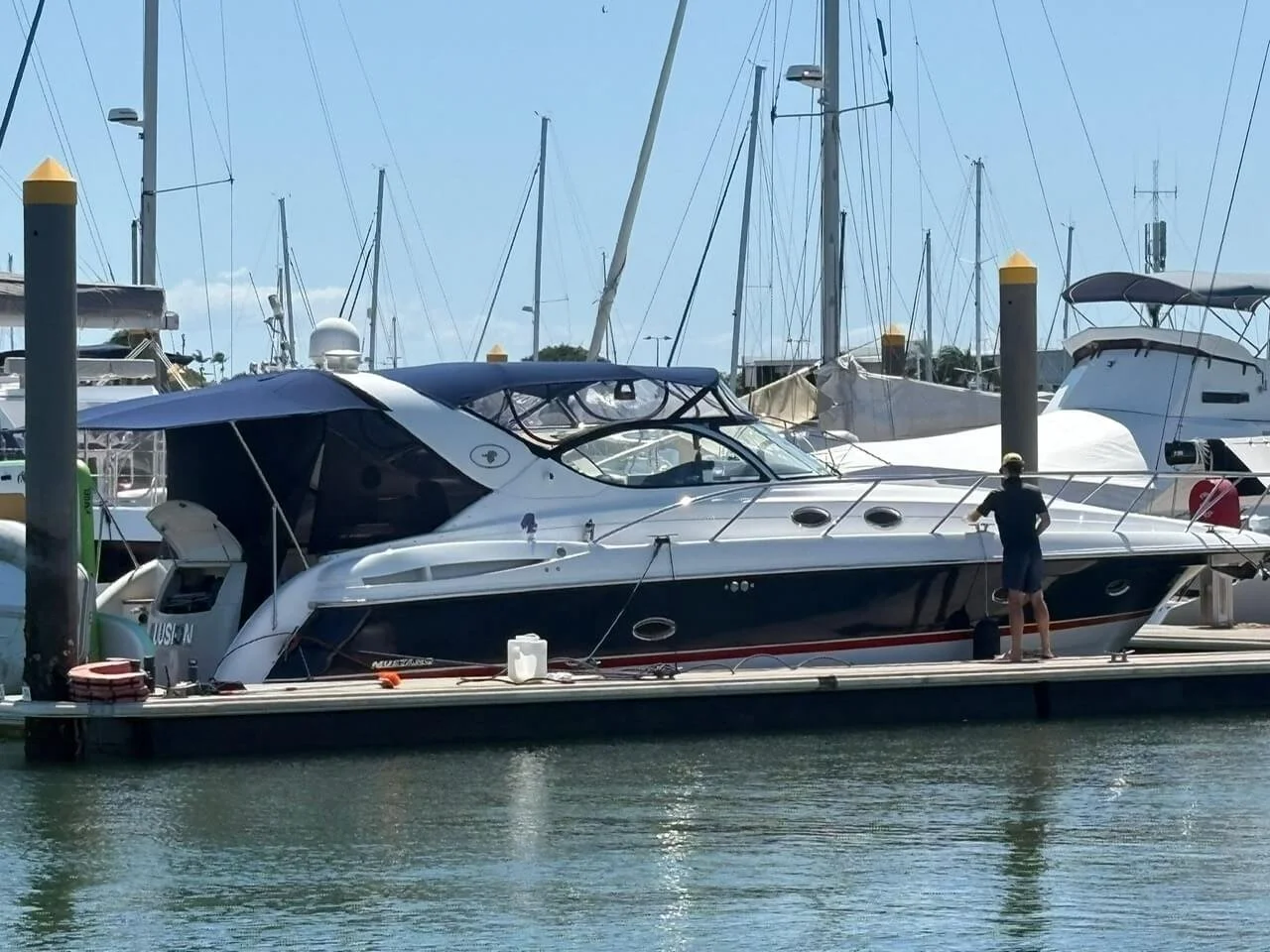 A person standing on a dock next to a large white and black yacht in a marina, with other boats and sailboats in the background under a blue sky.