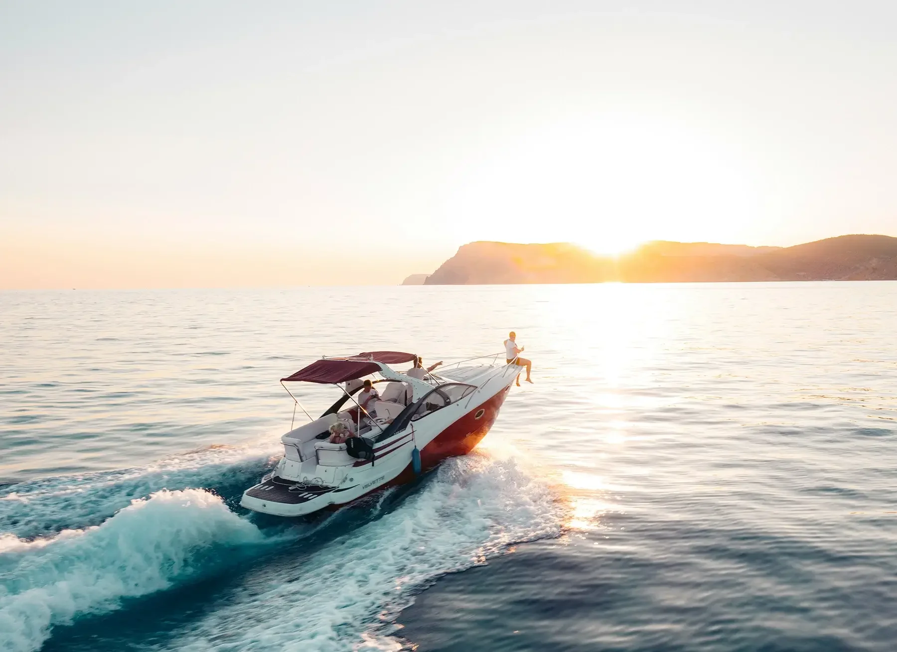 A motorboat speeding on the water during sunset with two people aboard, one sitting at the bow and another at the stern. The sun is setting behind a distant landmass, casting a golden glow over the scene.