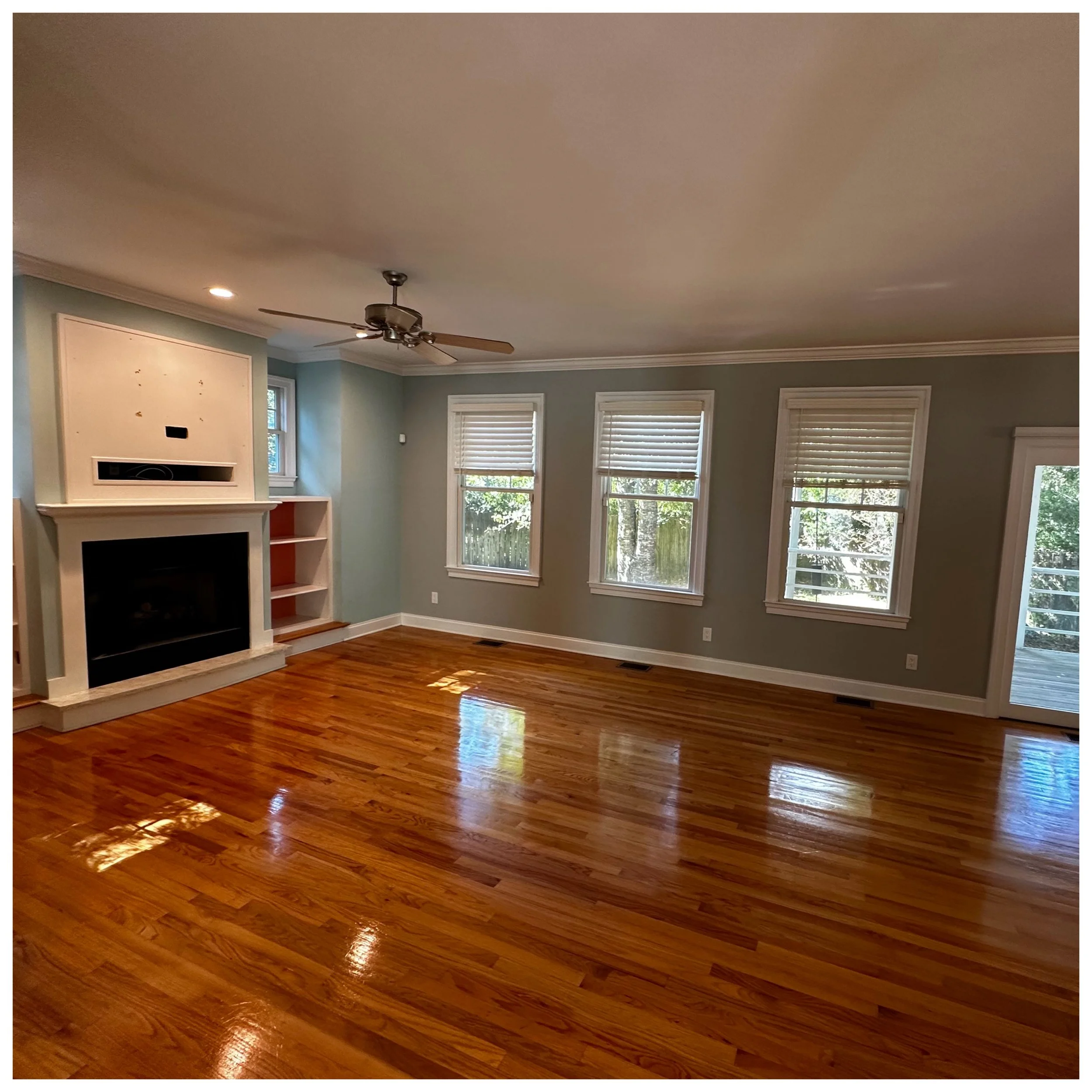 Empty living room with hardwood floors, three windows with closed blinds, a ceiling fan, and a fireplace with built-in shelves.
