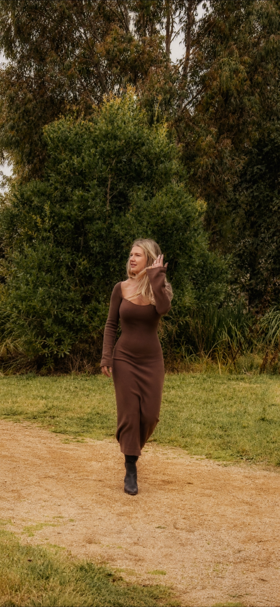 A woman with long blonde hair walking on a dirt path in a park, wearing a brown long-sleeve dress and black boots, with green trees and bushes in the background.