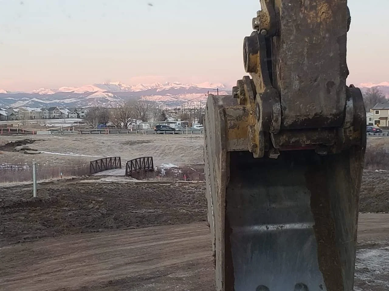 Close-up of a large excavator bucket at a construction site with snow-capped mountains in the background.