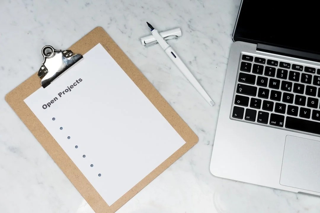 Clipboard with a paper titled 'Open Projects' on a white marble surface beside a silver laptop, a white pen, and a black marker.