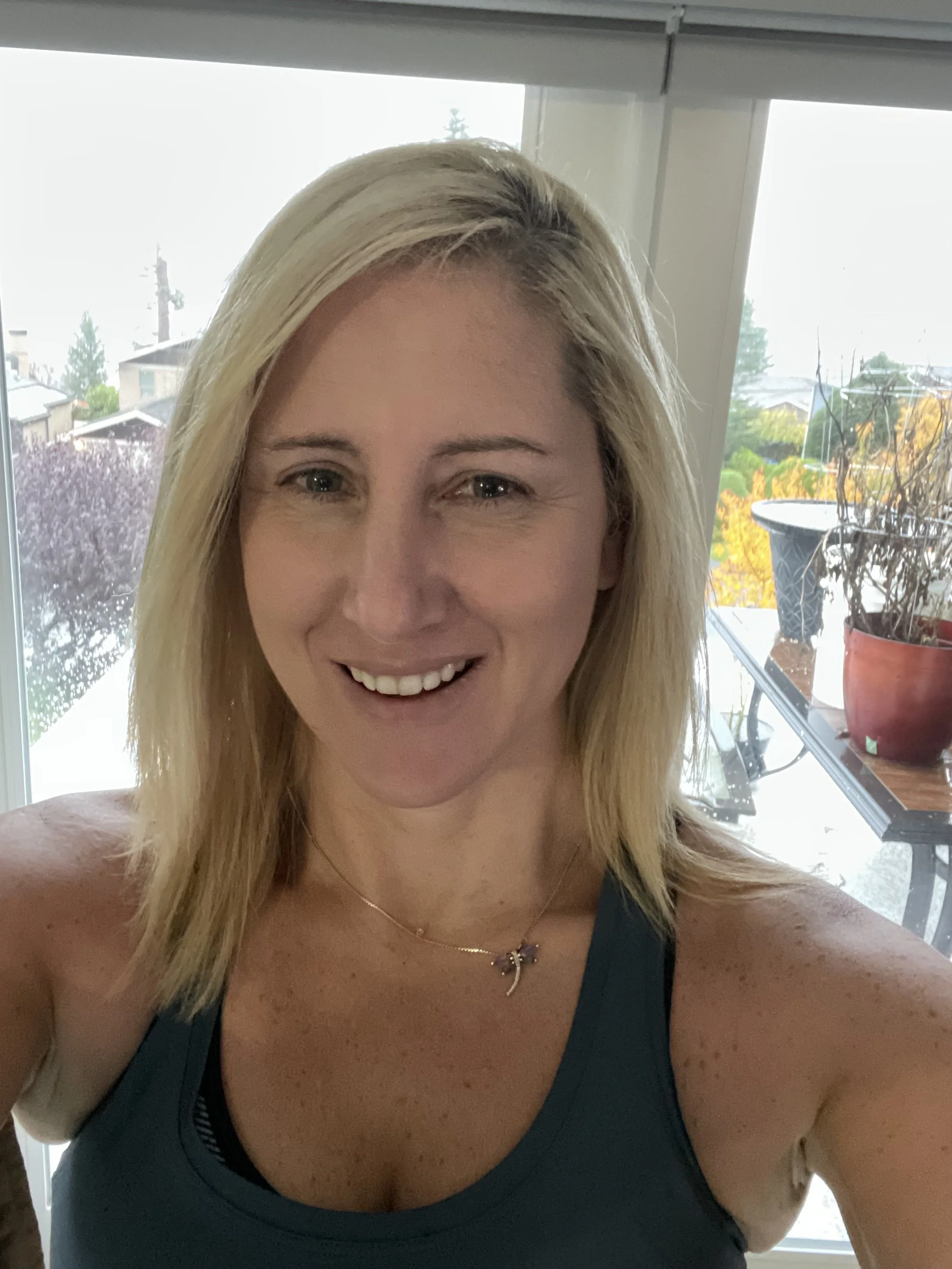 A smiling blonde woman taking a selfie indoors, with a window showing outdoor trees and patio furniture in the background.