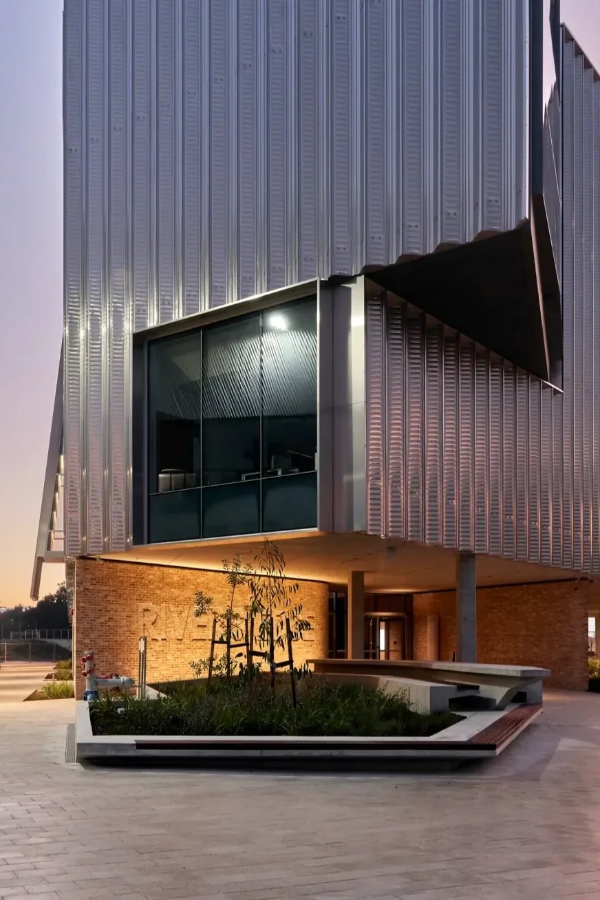 Modern building exterior with metal siding, large glass window, brick base, and small landscaped area with plants and a bench.