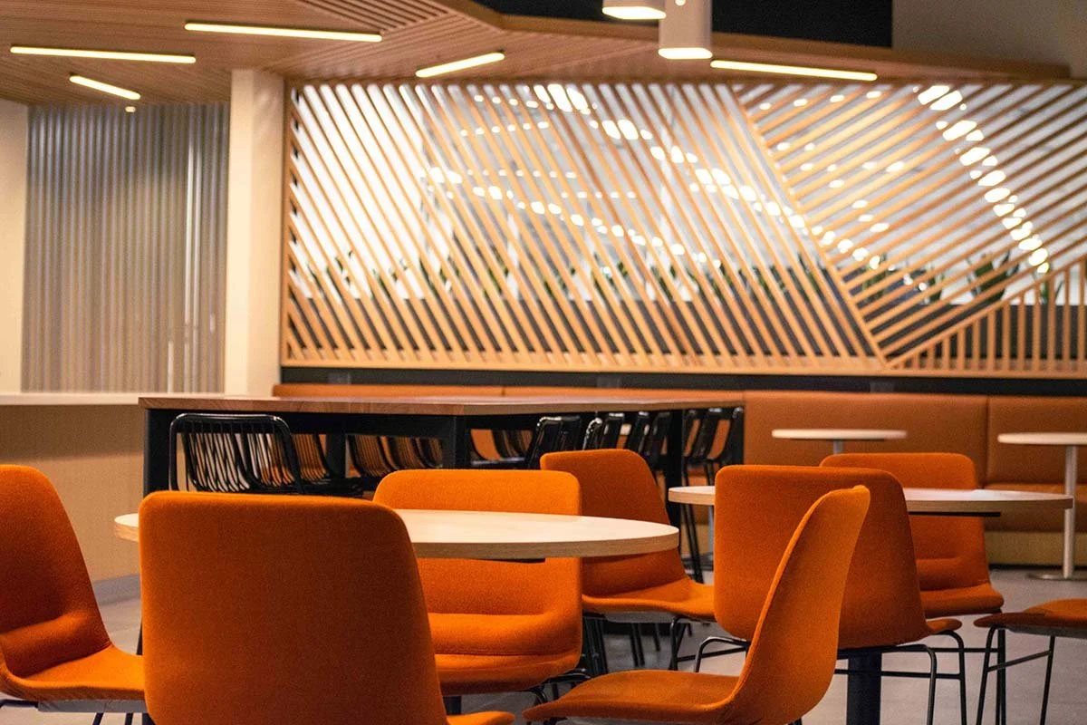 Empty seating area in a modern restaurant or café, with orange upholstered chairs and white round tables, wooden lattice wall, and soft lighting.
