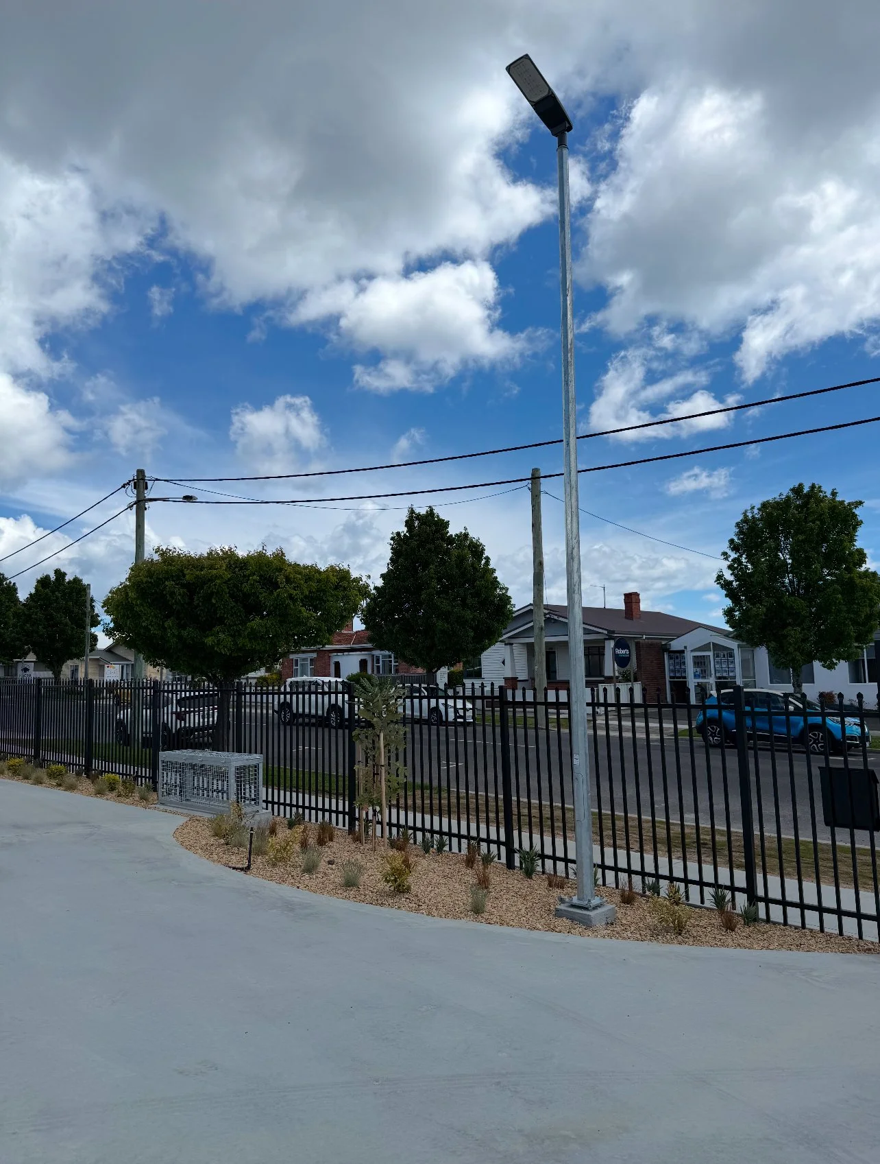 Residential neighborhood street with a black metal fence, trees, parked cars, a street lamp, and a partly cloudy sky.