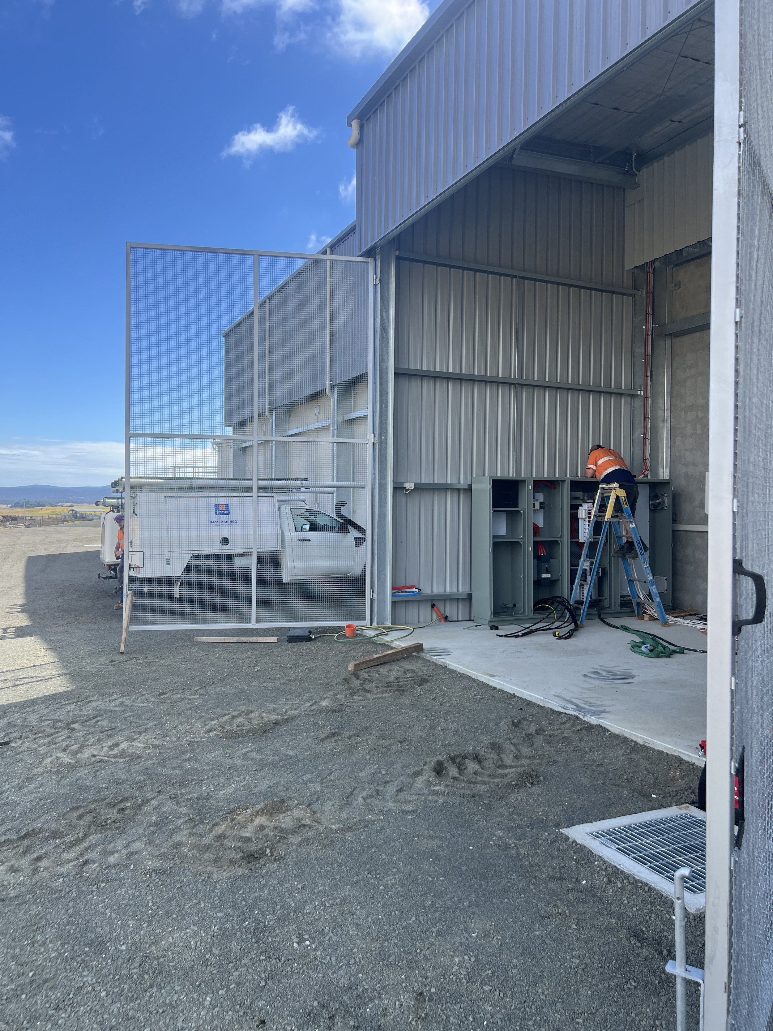 A worker in an orange safety vest standing on a ladder inside a metal storage building, working on electrical or technical equipment.