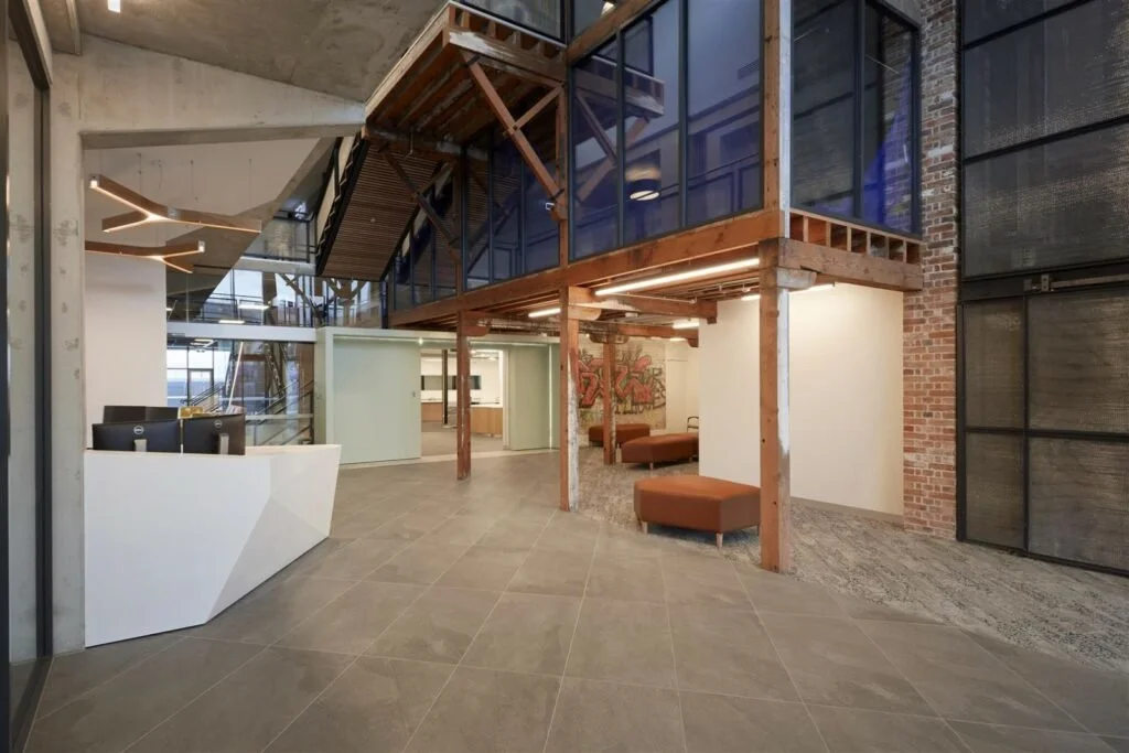 Modern office lobby with a reception desk, orange seating, and an open second floor with glass walls and wooden supports.