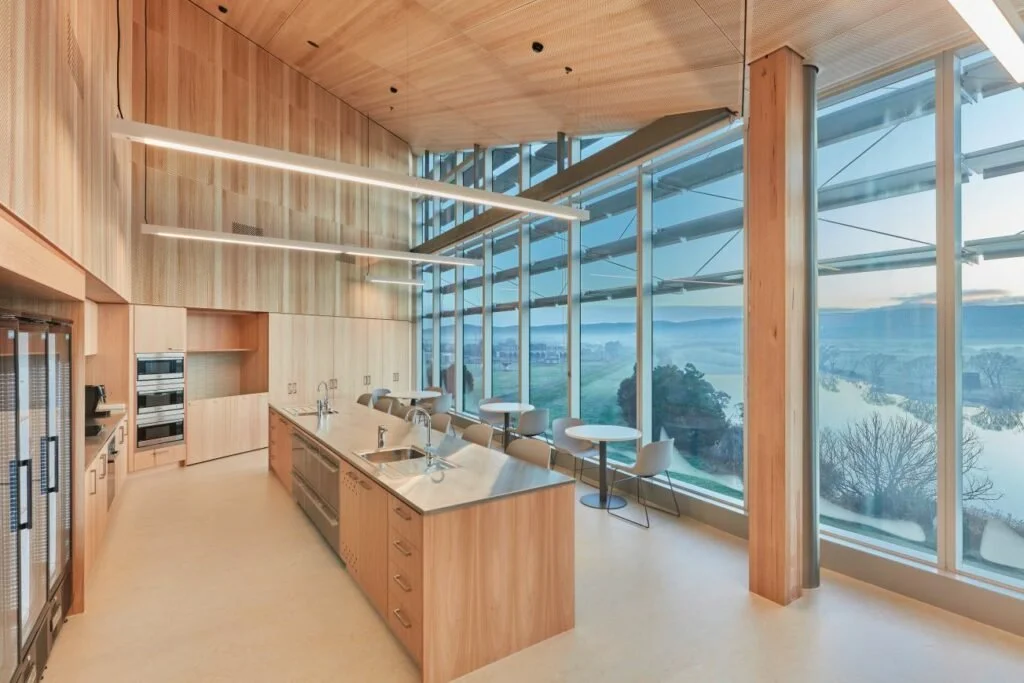 Modern kitchen with a wooden finish, large glass windows showing a scenic landscape, and minimalistic furniture.