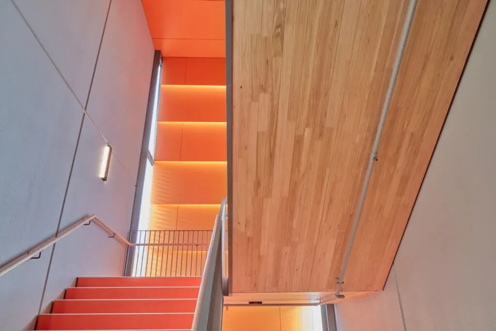 Interior view of a modern building with a staircase, wooden wall, gray ceiling, and orange accents along the stairs and wall panels.
