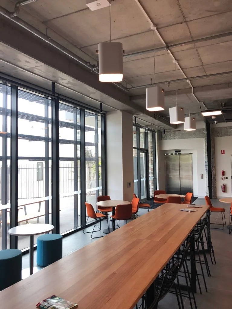 Interior of a modern café or lounge area with a large wooden table, orange chairs, small round tables near large glass windows, pendant lights, and an elevator in the background.