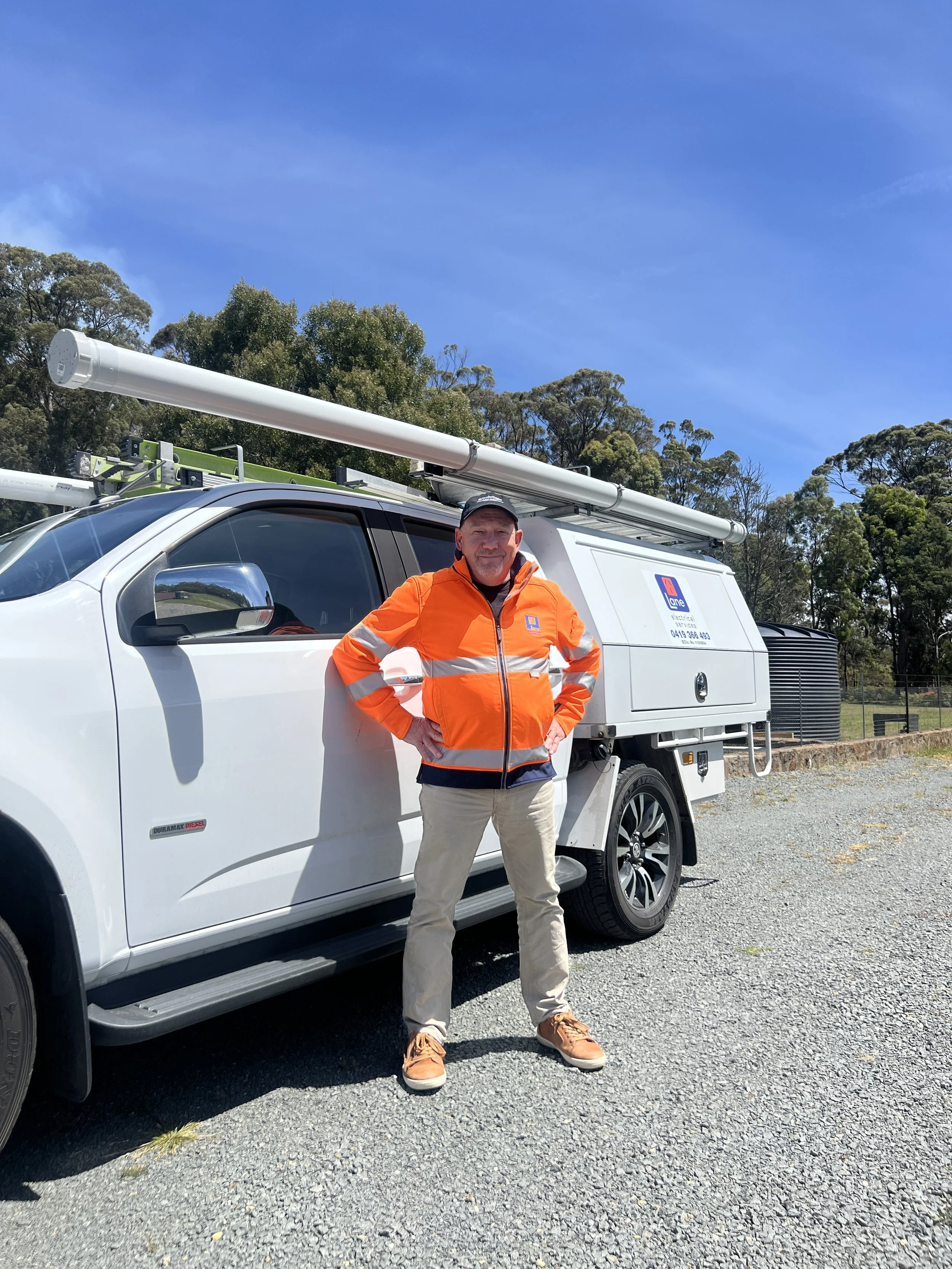 A man standing next to a white utility truck with equipment on the side, outdoors on a sunny day with trees and a partly clear sky in the background.