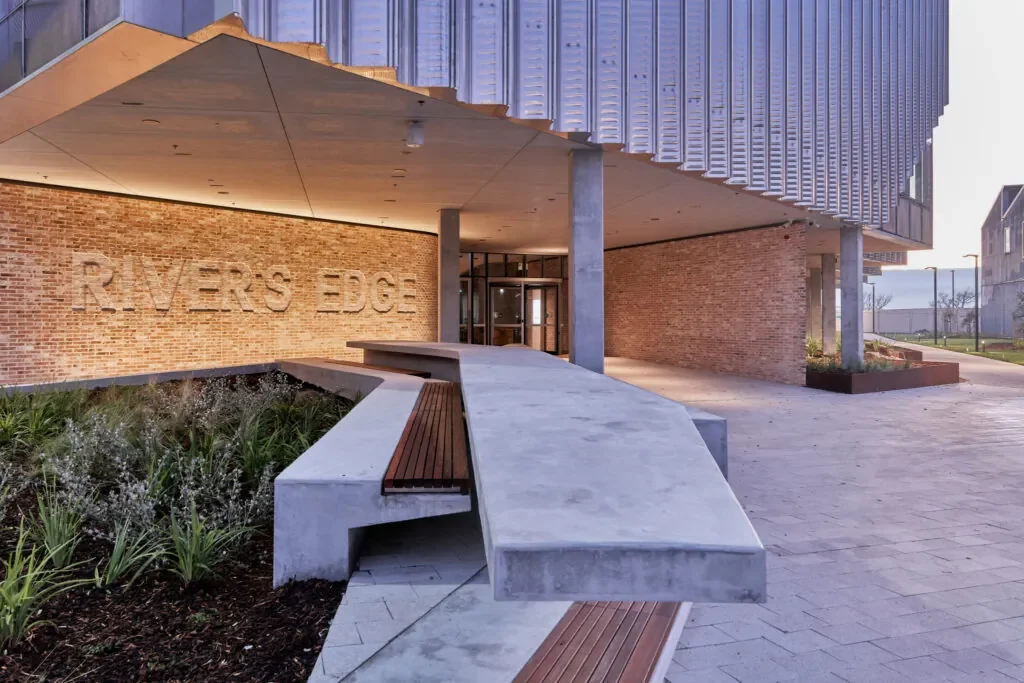 Exterior of a modern building with a brick facade and the words 'RIVERS EDGE' engraved on the wall. A curved concrete bench with wooden slats is in the foreground, surrounded by landscaped plants. The building has metal siding and large glass doors.