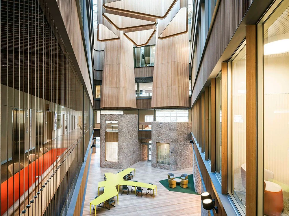 Interior view of a modern building with wooden and brick accents, large windows, and colorful furniture including yellow tables and green seating.