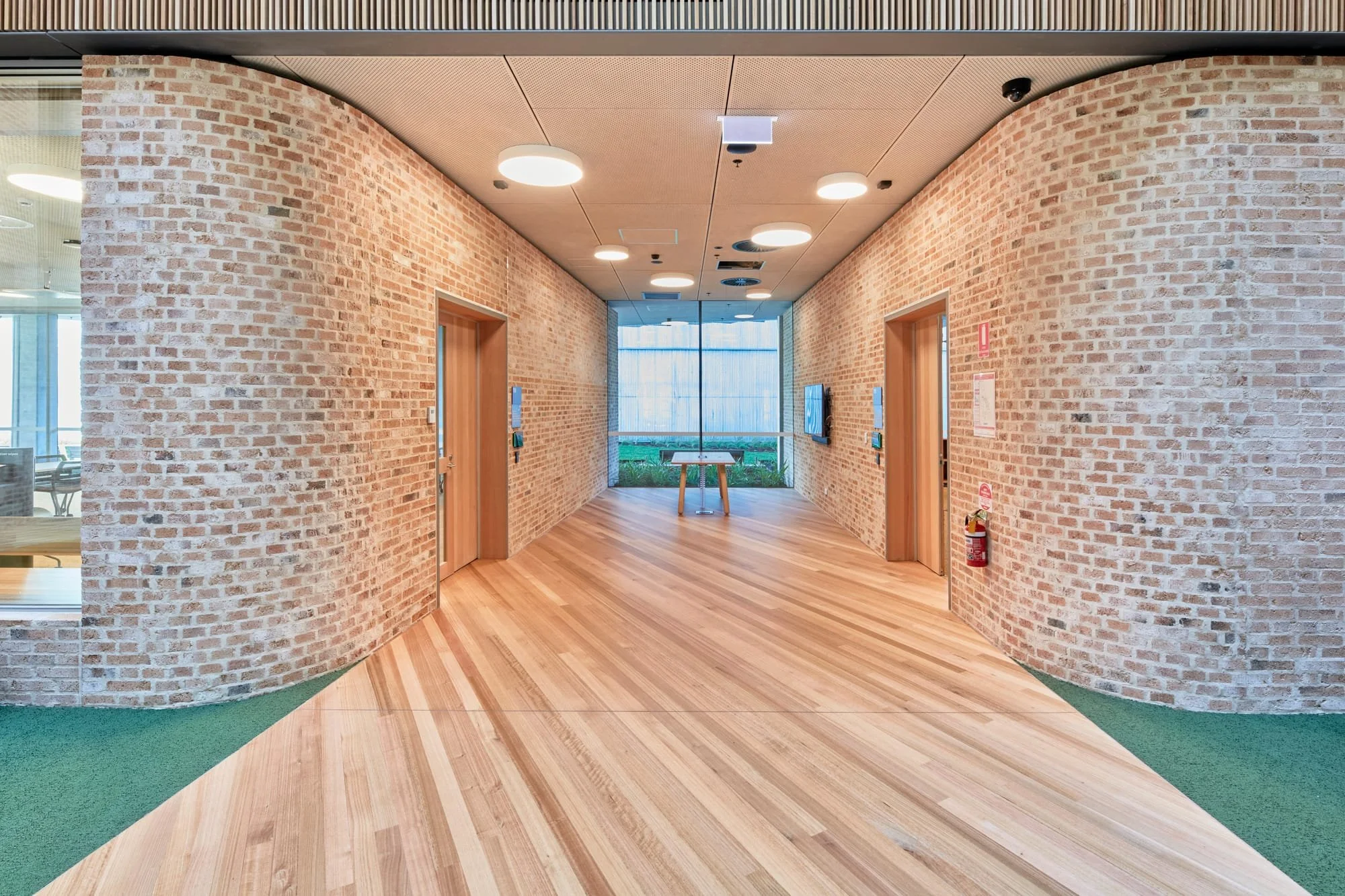 Interior hallway with brick walls and wooden floors, two elevator doors, a small window at the end with a table, ceiling lights, and a fire extinguisher on the right wall.