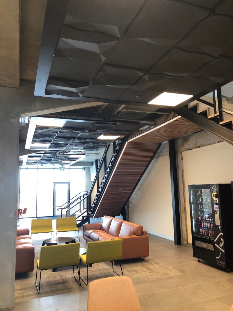 Modern lobby with colorful chairs and a staircase leading to an upper floor, vending machine, and large windows in the background.