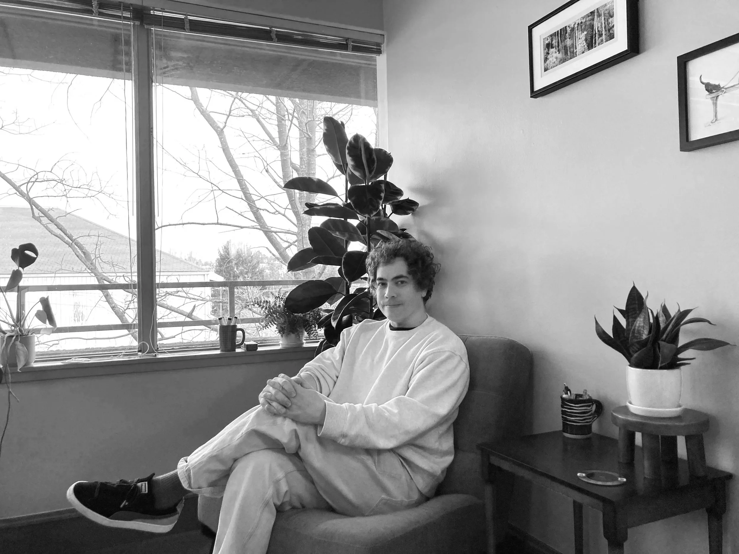 A young person with curly hair sitting on a couch in a living room, surrounded by indoor plants near a large window with a view of trees and rooftops, in black and white.