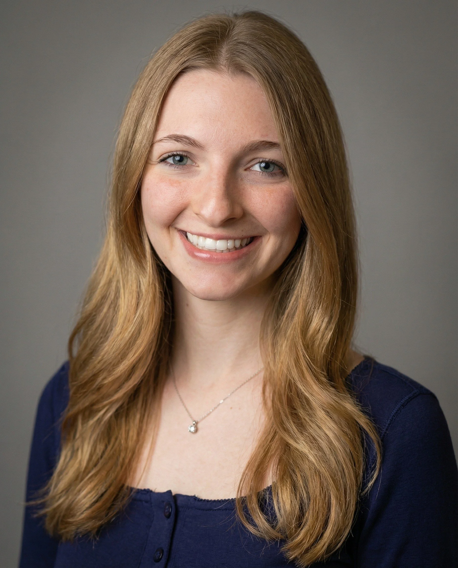 A young woman with long, wavy red hair and blue eyes smiling, wearing a navy top and a delicate silver necklace, against a plain gray background.