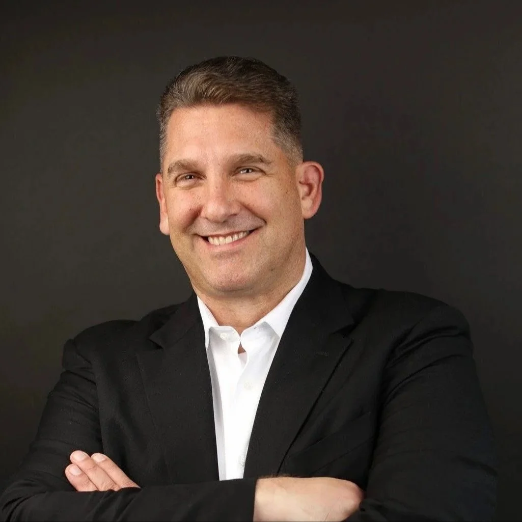 A smiling man in a black suit with arms crossed against a dark background.