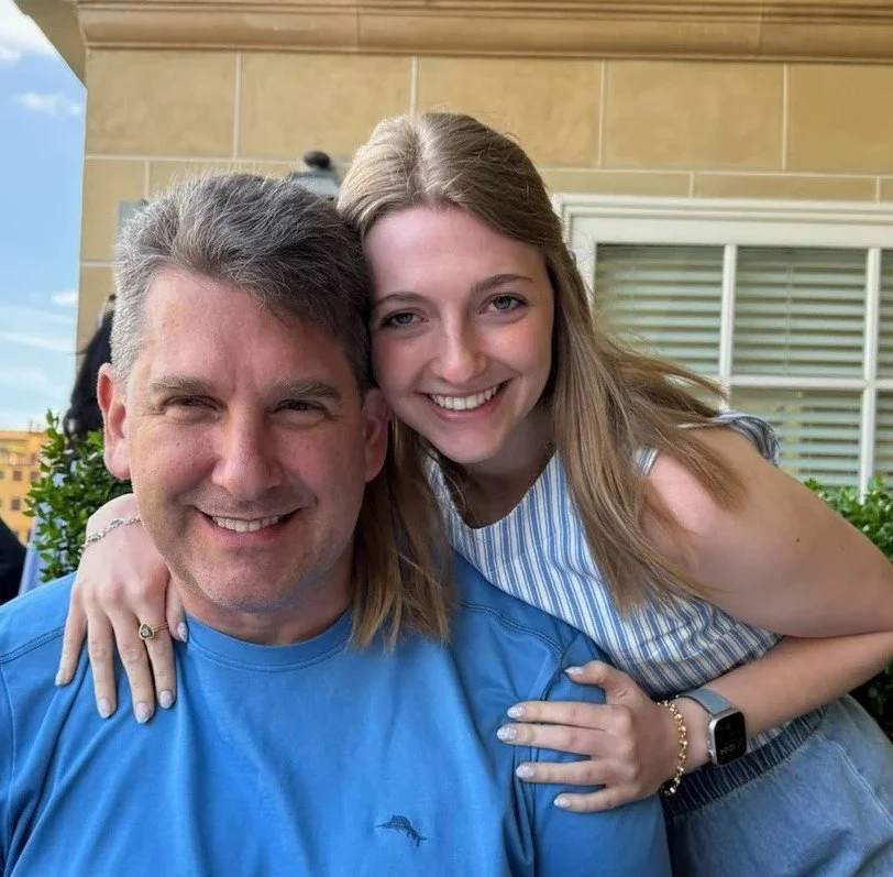 A smiling father and daughter posing outdoors, with the daughter hugging the father from the side, both wearing casual clothing, against a building background.