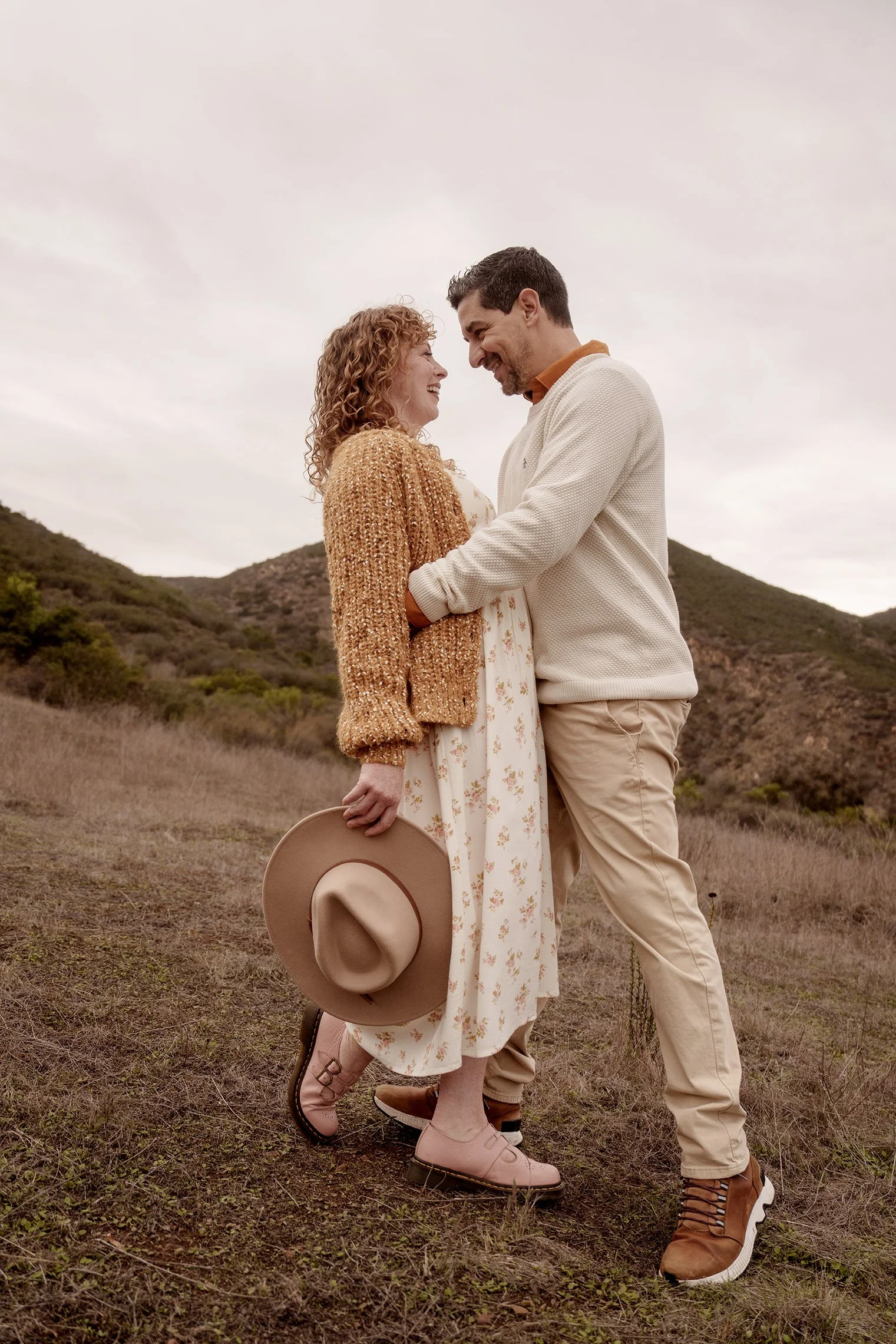 A couple standing close together on a grassy hillside, smiling and looking into each other's eyes, with mountains in the background and an overcast sky.