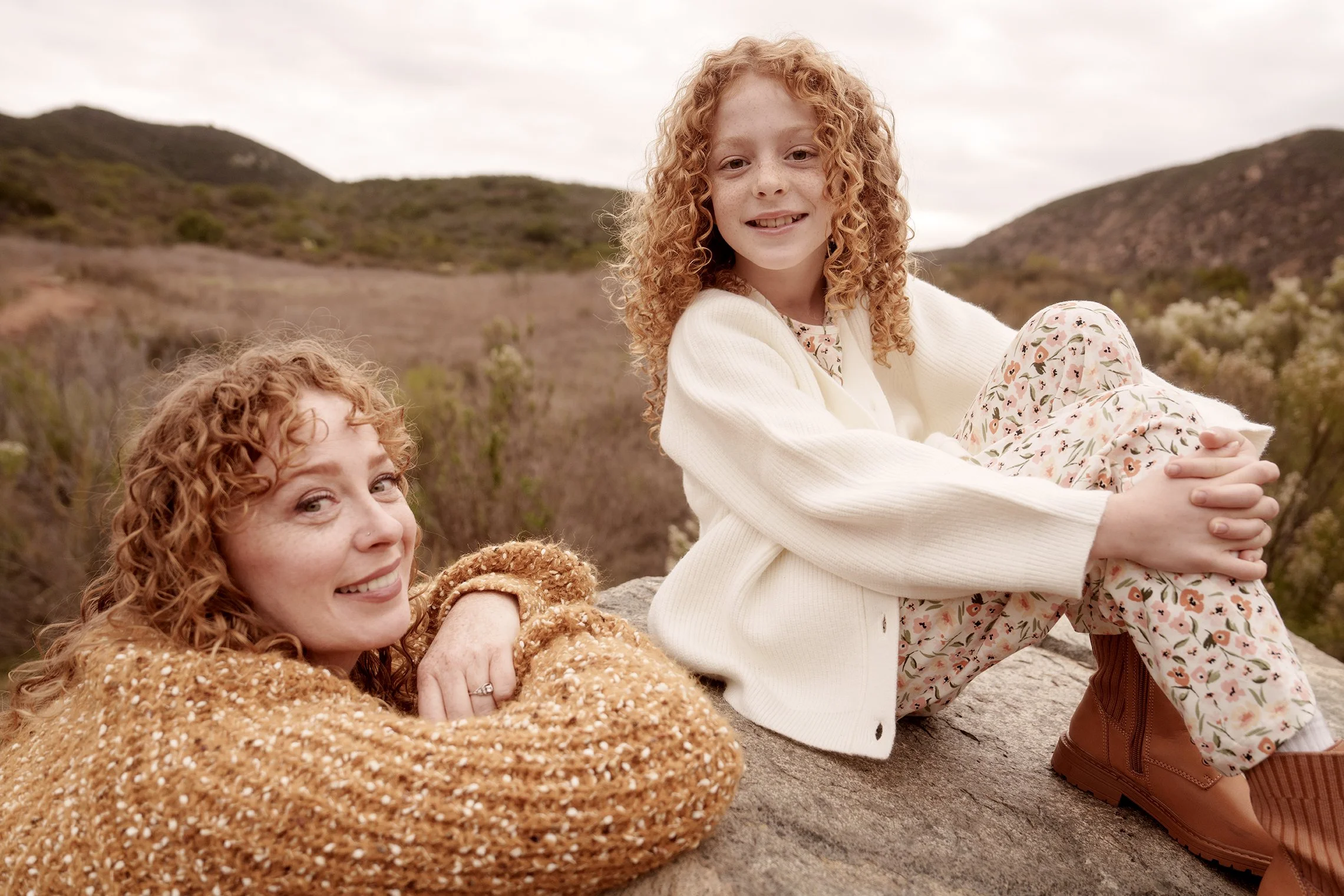 A woman with curly red hair and a young girl with curly red hair sitting outdoors on a rock in a hilly landscape. The woman is smiling and resting her head on her arm, wearing a brown textured sweater. The girl is sitting cross-legged, smiling at the