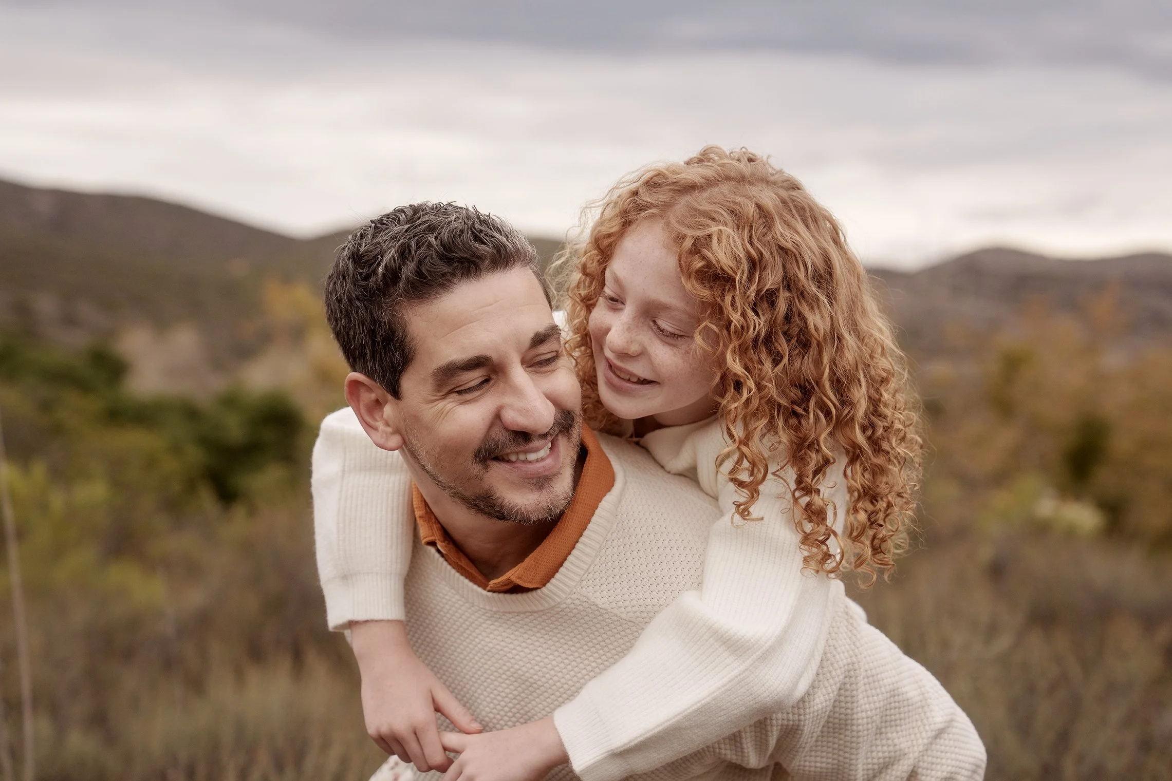 A father and daughter outdoors, with the daughter riding on the father's back, smiling and looking at each other, with mountains and cloudy sky in the background.