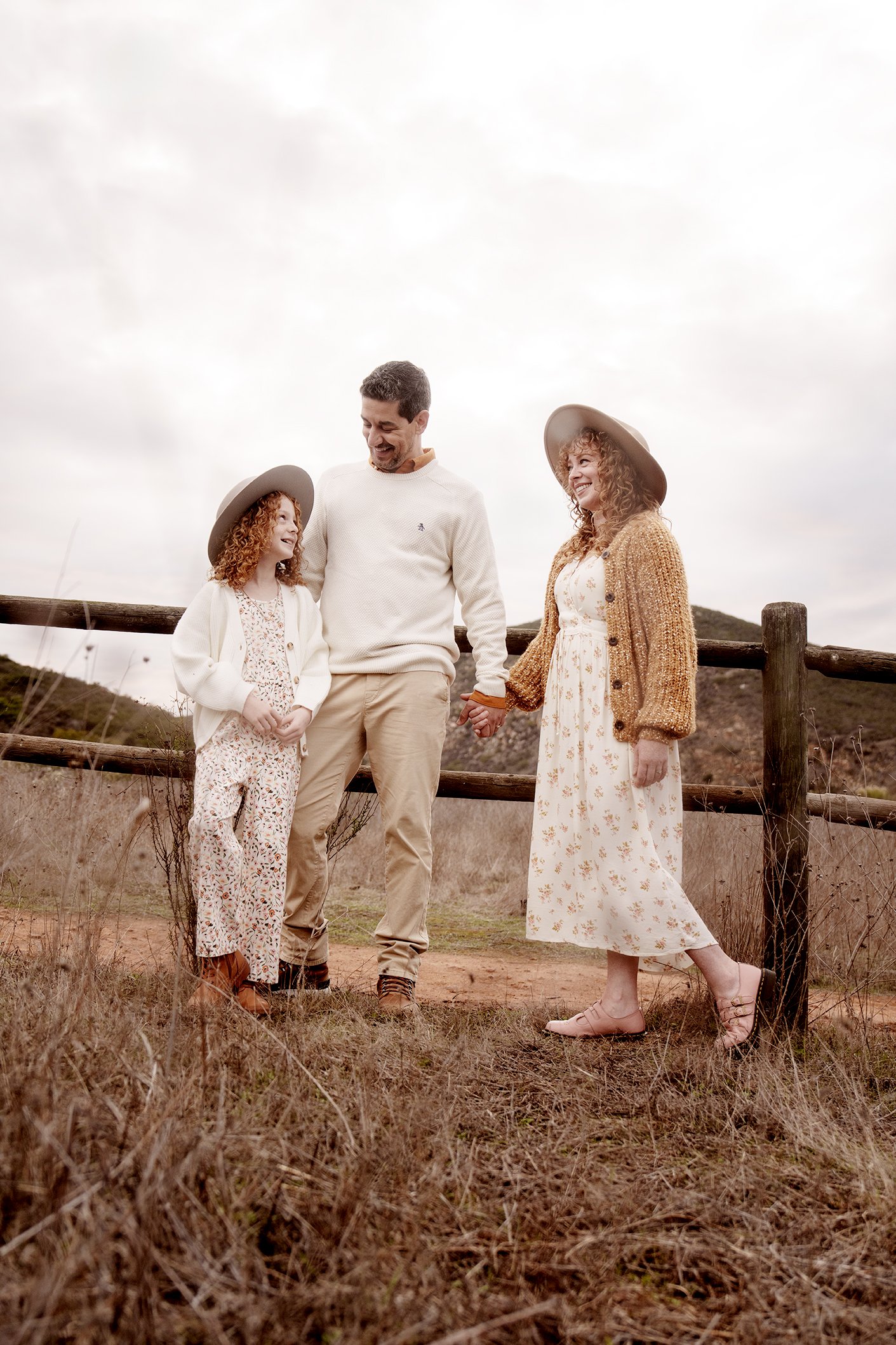 A family of three standing outdoors on a dirt path near a wooden fence, holding hands and smiling at each other.