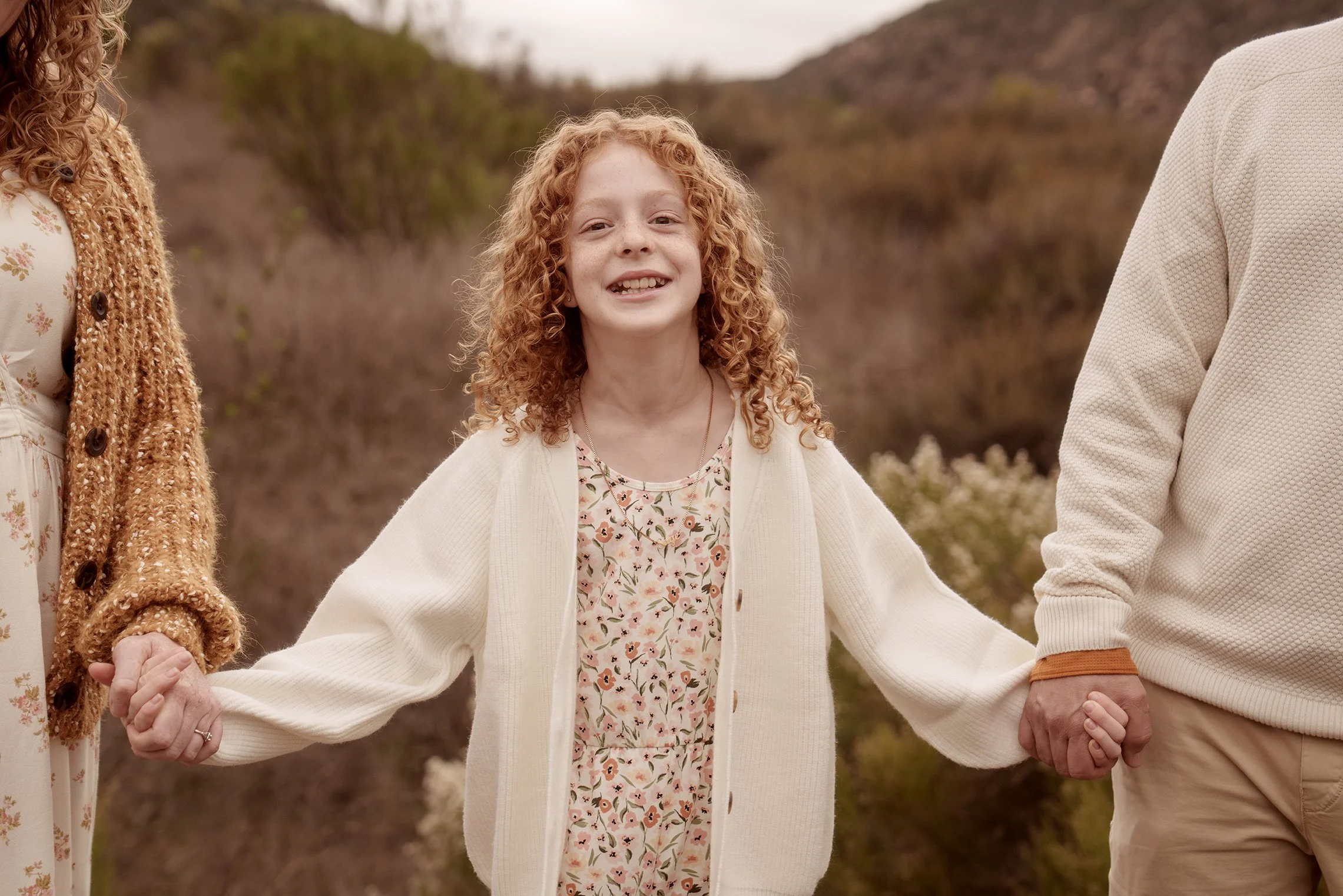 A young girl with curly red hair smiling and holding hands with two adults outdoors.