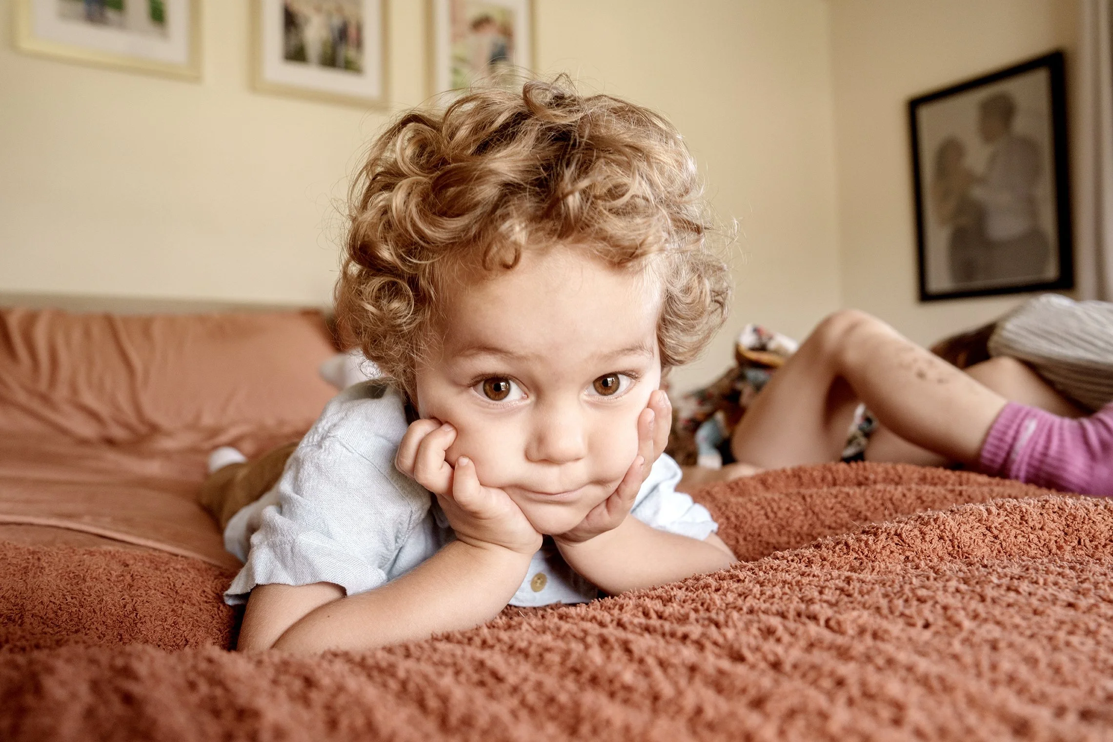 A young boy with curly red hair lying on a bed, resting his chin on his hands, looking at the camera with a curious expression. There are framed photos on the wall behind him.