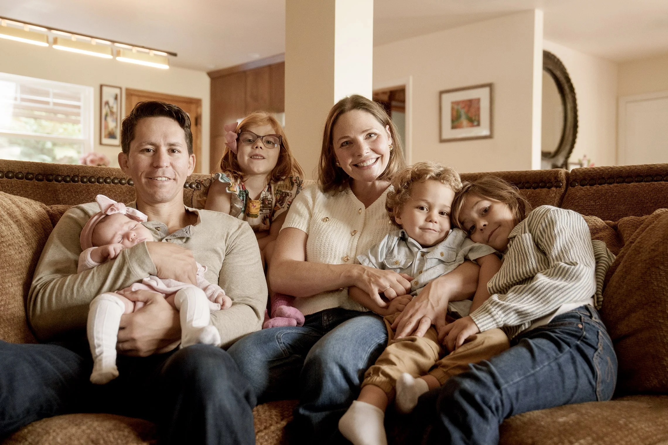 A happy family of seven sitting on a beige couch in a cozy living room, with a man, woman, and five children including an infant, with cheerful expressions.