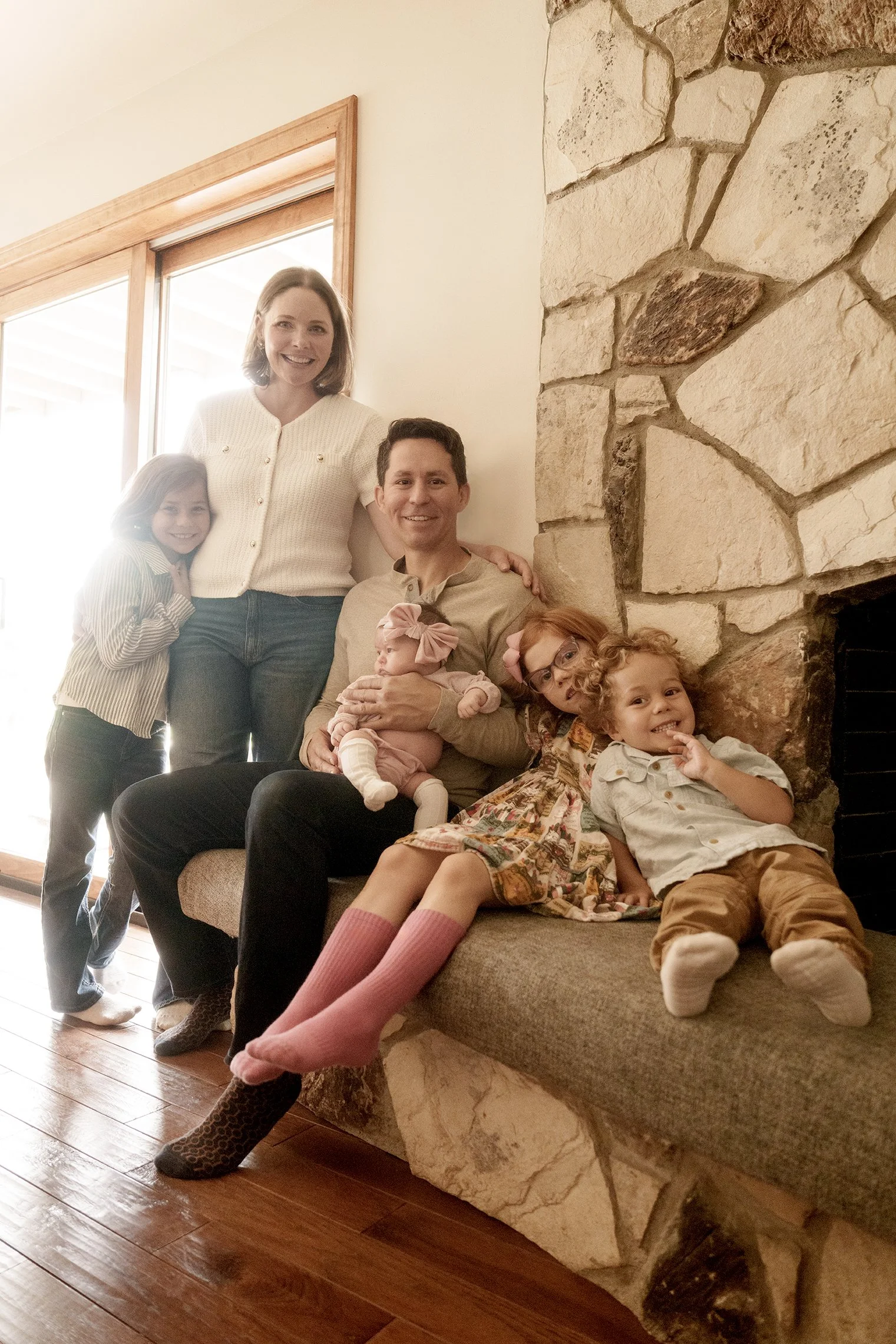 Family of seven sitting on a bench by a stone fireplace and a sliding glass door, smiling at the camera.