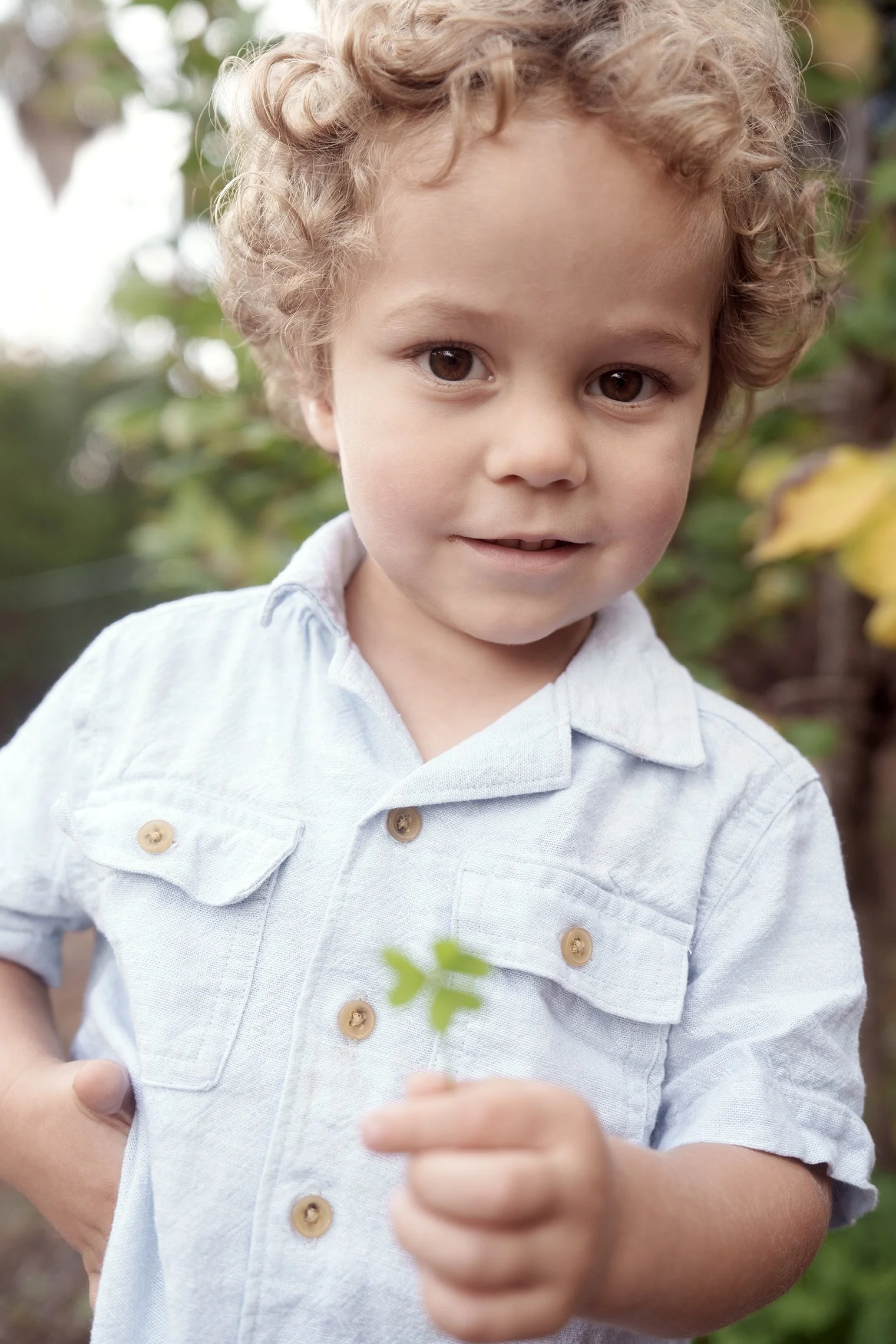 A young boy with curly blonde hair and brown eyes holding a small green leaf in his hand outdoors, smiling slightly with a natural background of foliage.