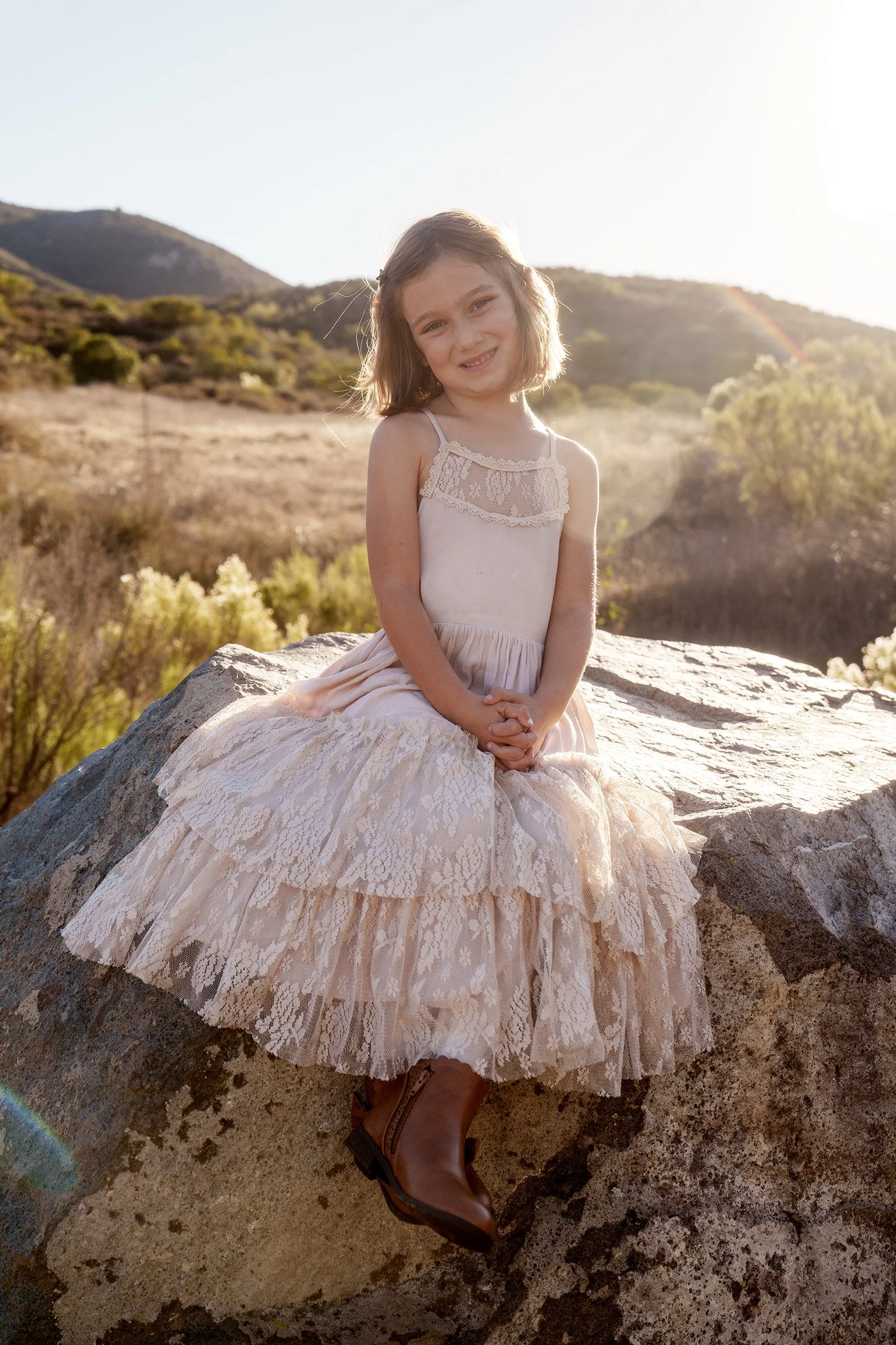 A young girl sitting on a large rock outdoors, wearing a light-colored dress with lace details and brown boots, smiling at the camera with a mountainous landscape and bright sunlight in the background.