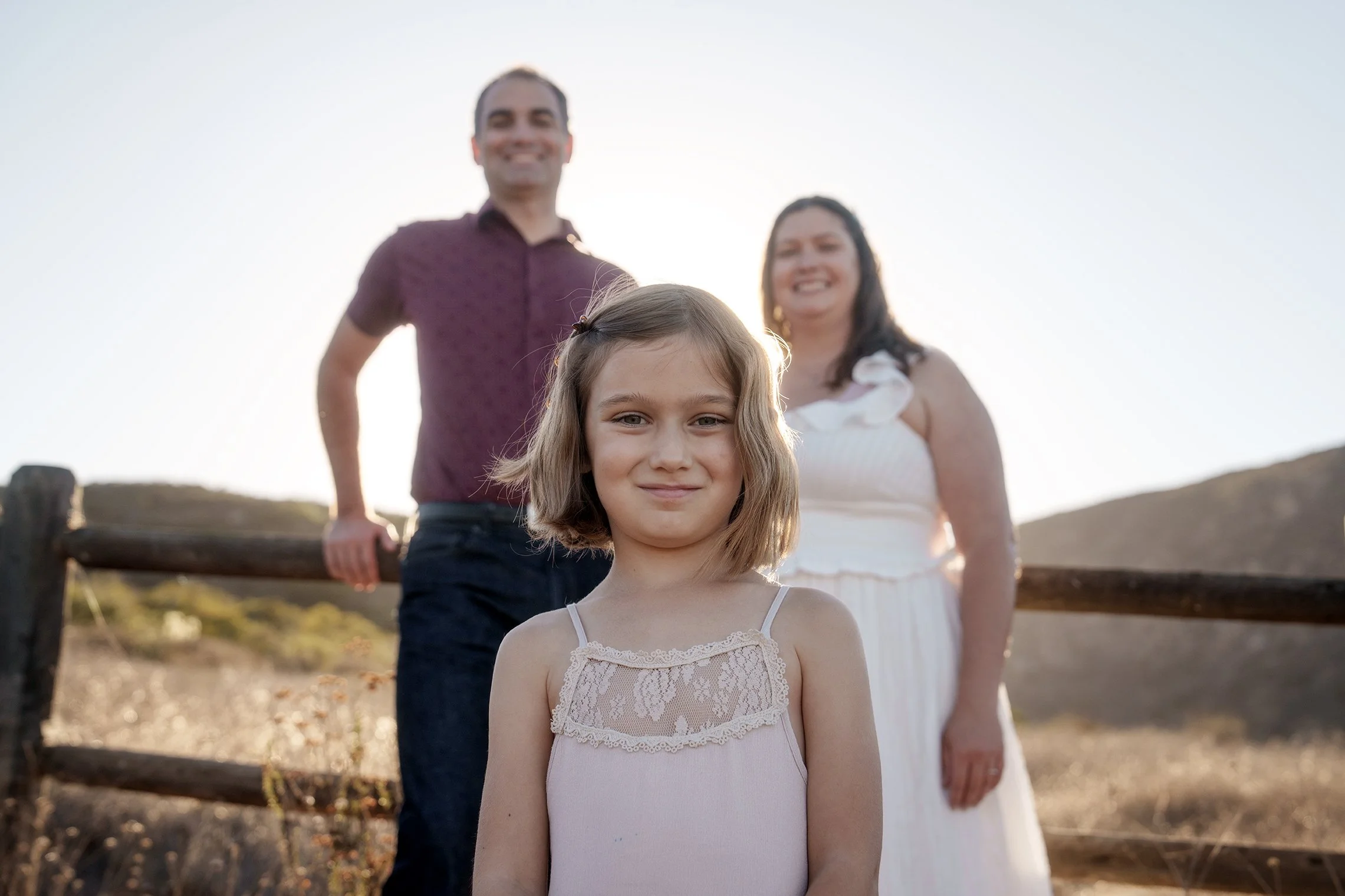 A young girl with shoulder-length blonde hair standing outdoors in the foreground, with a man and woman smiling behind her, near a wooden fence during sunset.