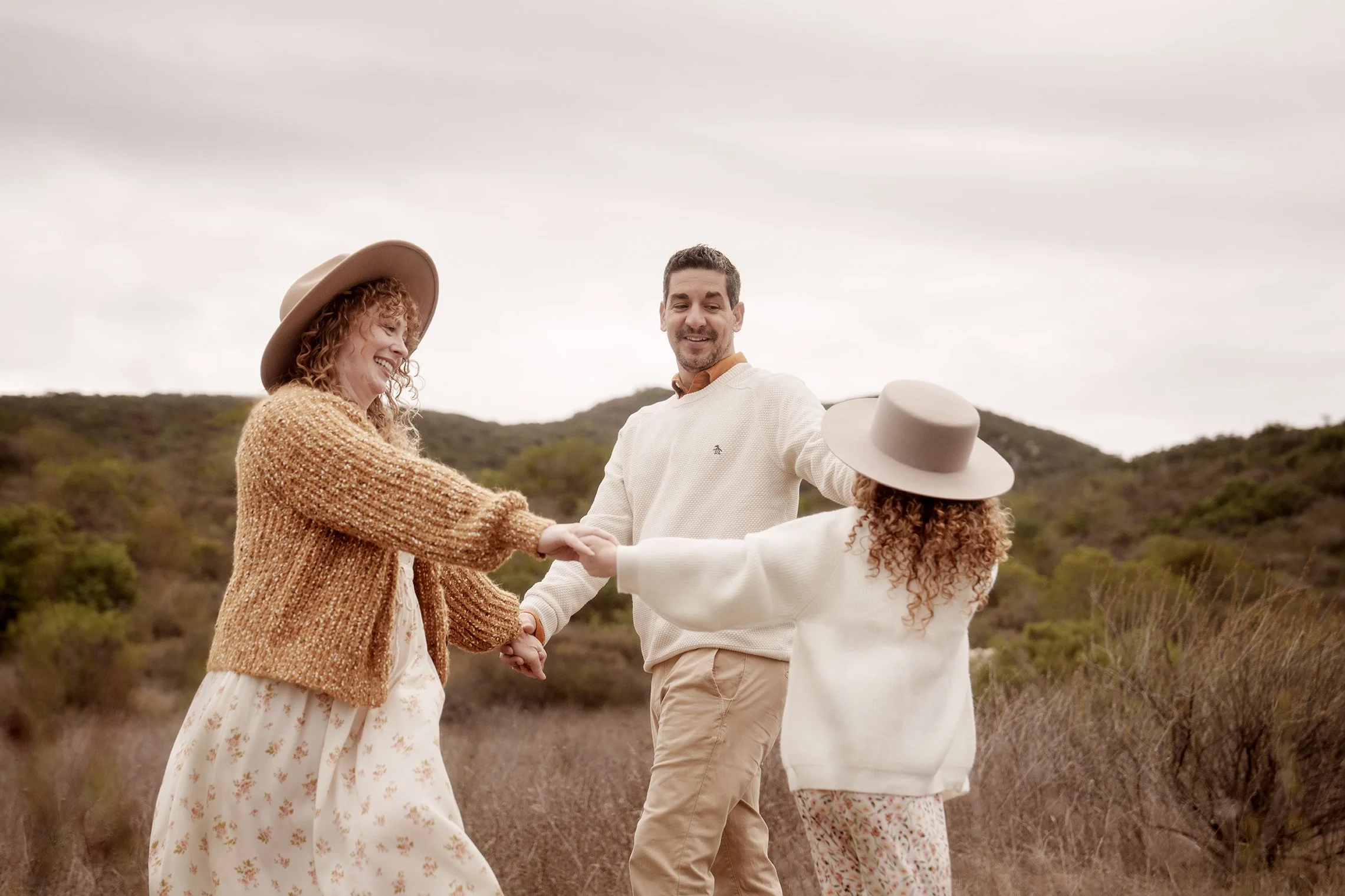 Three people, one man and two women, holding hands and dancing outdoors in a hilly landscape with bushes and trees, under a cloudy sky.