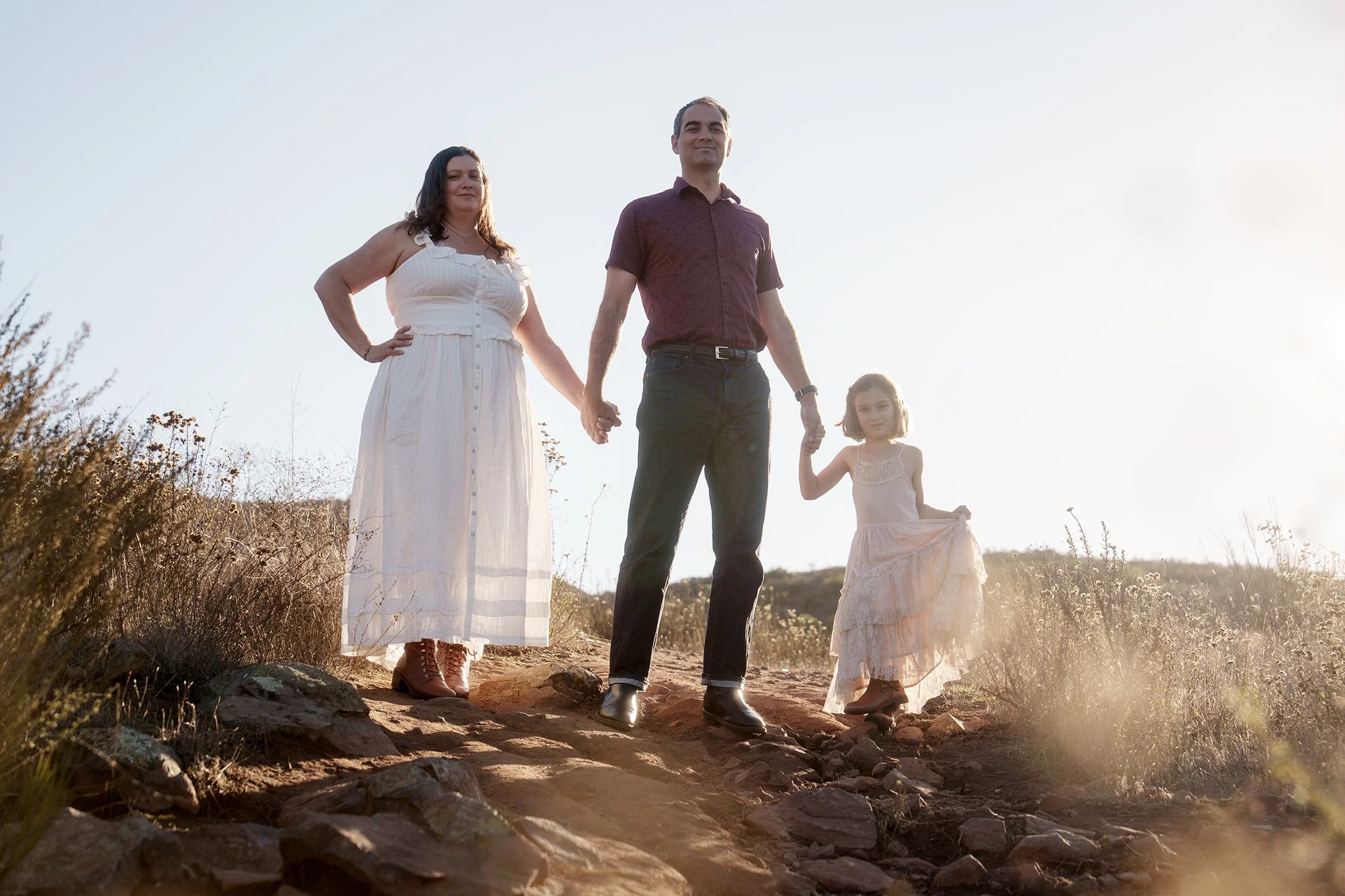 A family of three holding hands outdoors in a sunlit, rocky, grassy area. The woman is wearing a white dress and boots, the man is in a maroon shirt and dark pants, and the girl is in a light-colored dress and boots.
