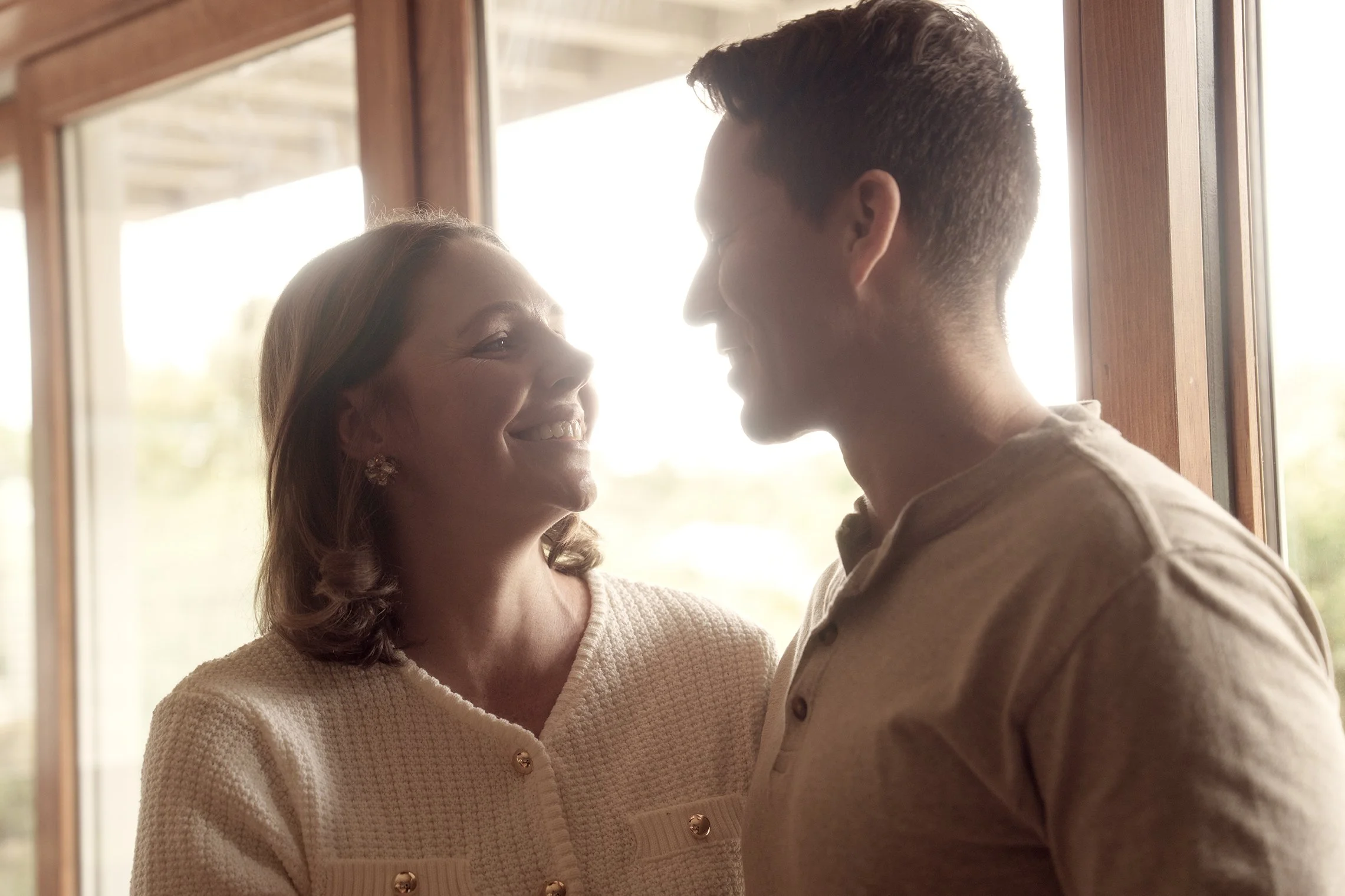 A woman and a man standing close to each other indoors, smiling and looking into each other's eyes at a window with natural light.