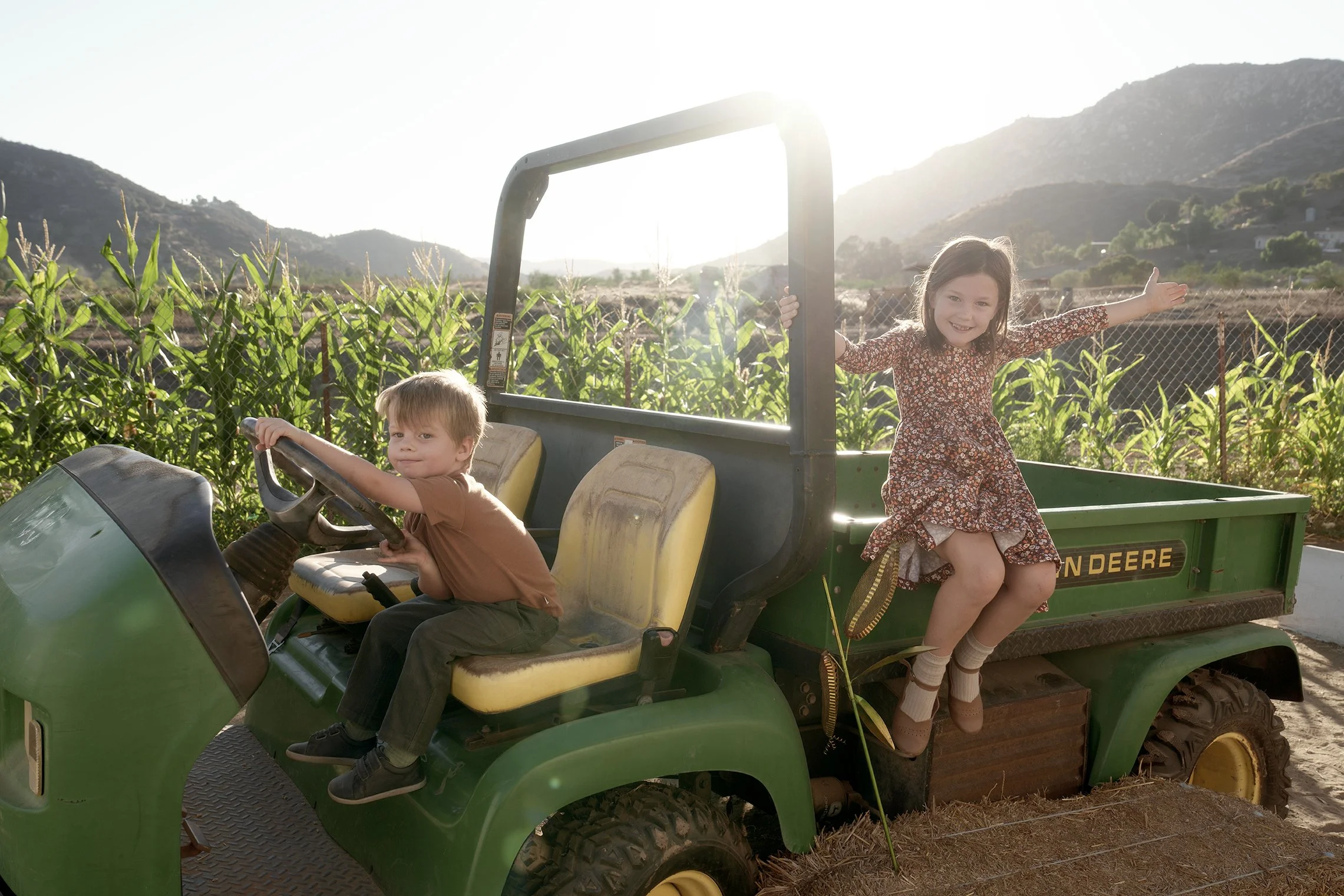 Two children, a boy and a girl, sitting on a green John Deere utility vehicle in a farm field with corn plants and mountains in the background, children smiling and enjoying the outdoors.
