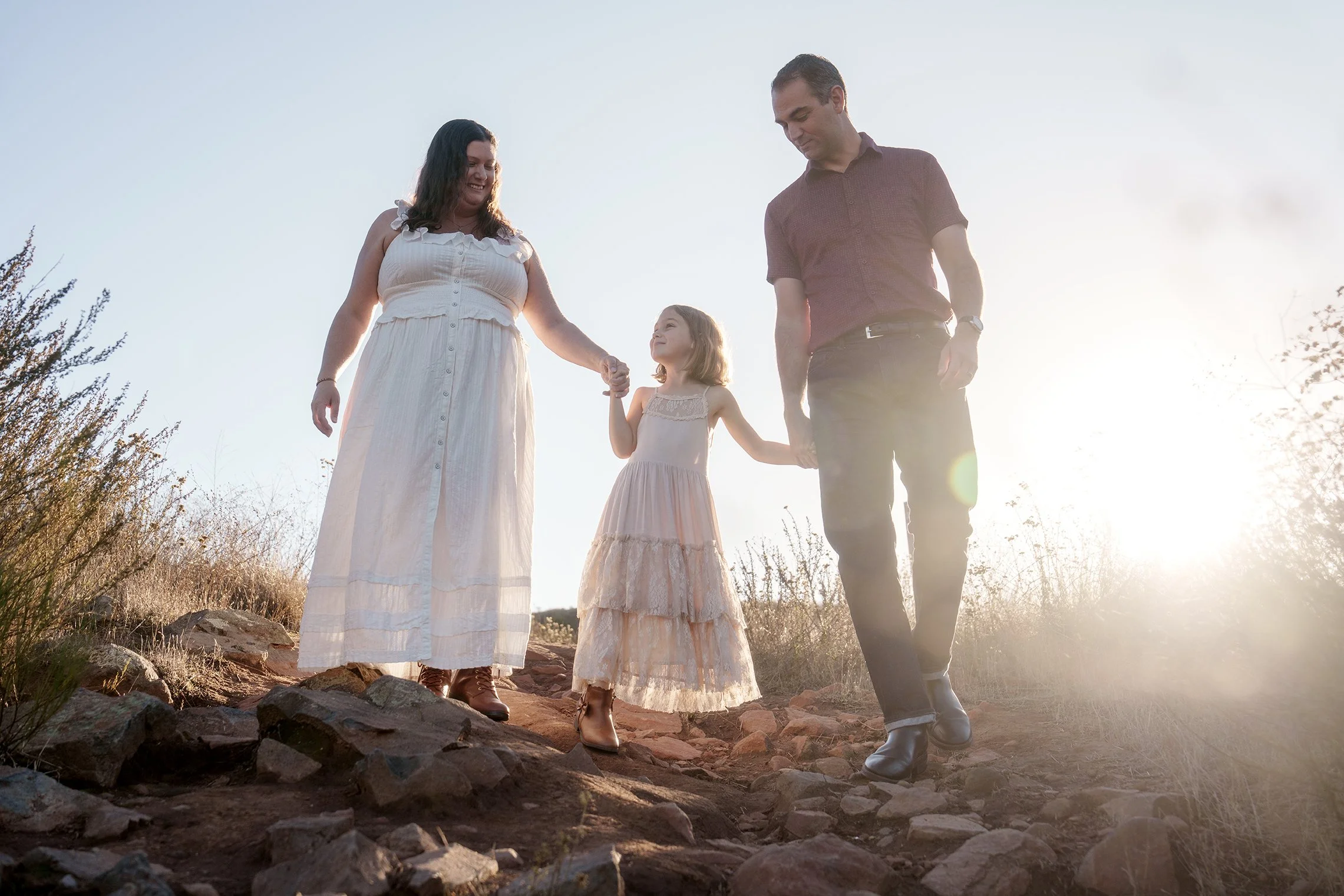 A family of three walking on a rocky trail outdoors during sunset, holding hands and smiling