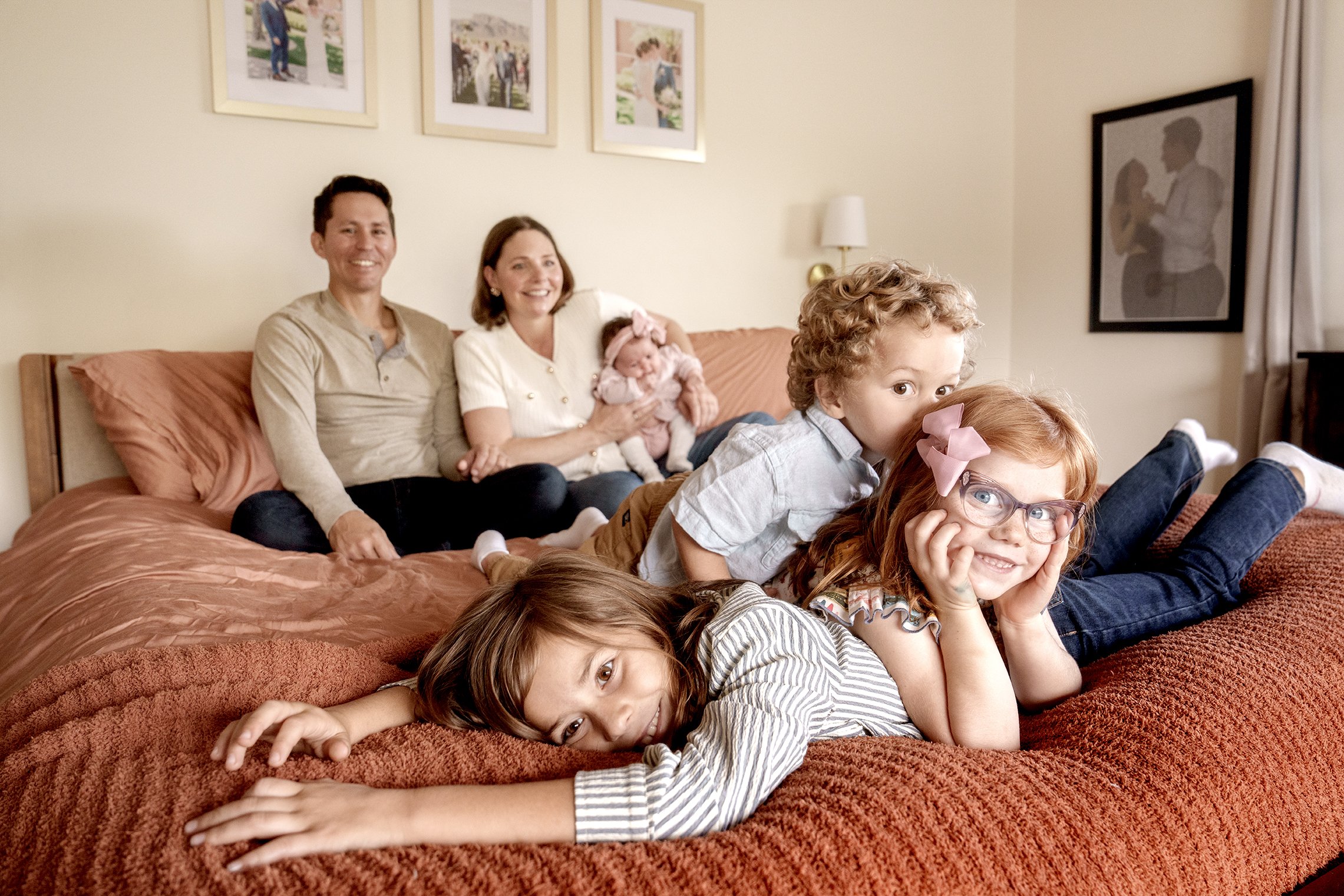 A family of six children and two adults lounging together on a bed in a cozy bedroom, with framed photos on the wall behind them.