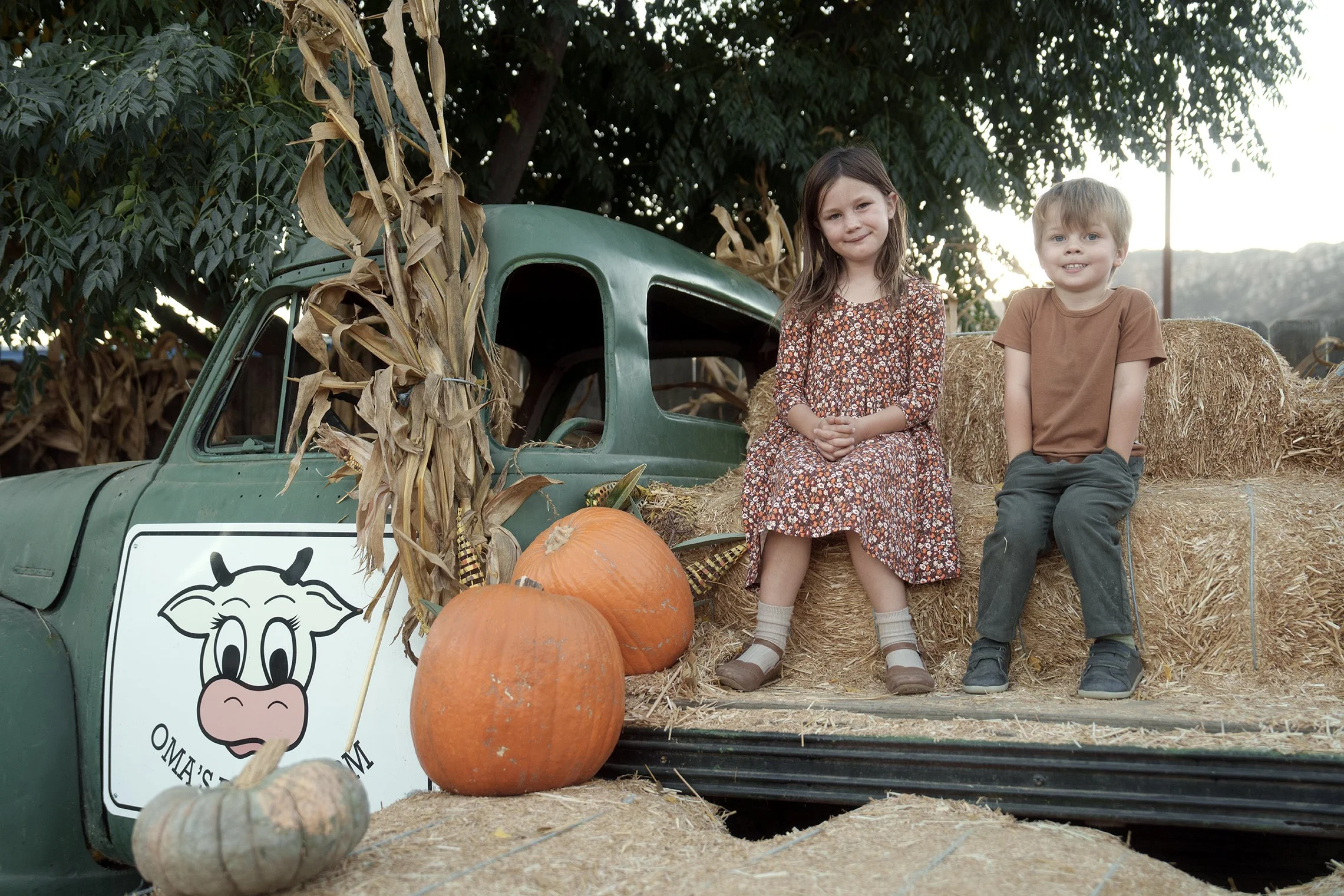 Two children, a girl in a floral dress and a boy in a brown shirt and gray pants, sitting on hay bales next to a decorated truck with pumpkins and a scarecrow on it, surrounded by corn stalks.