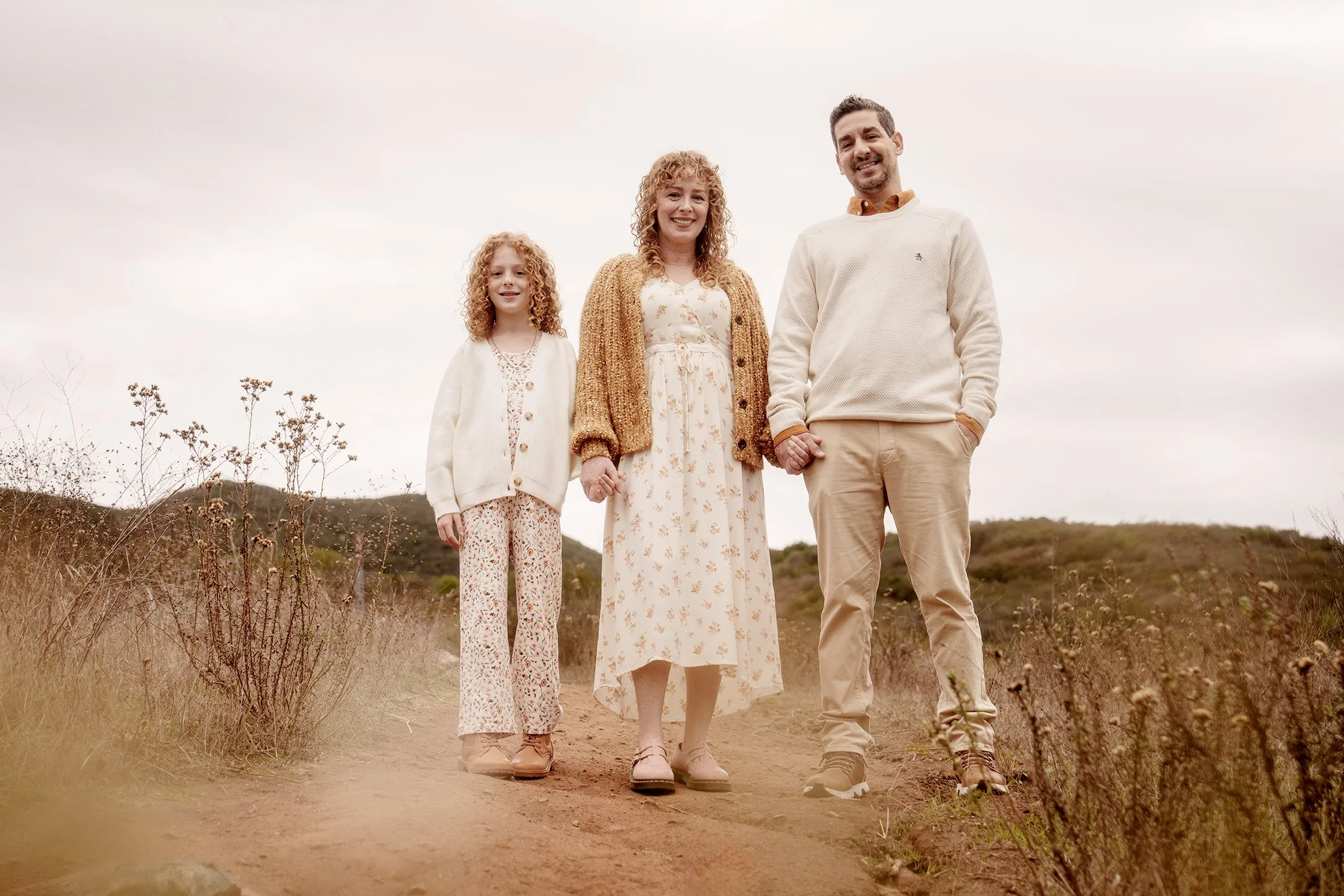 A family of four, consisting of a mother, father, and two daughters, standing on a dirt path outdoors in a natural setting with hills and dry vegetation in the background. They are holding hands, smiling, and dressed in casual, neutral-colored clothi