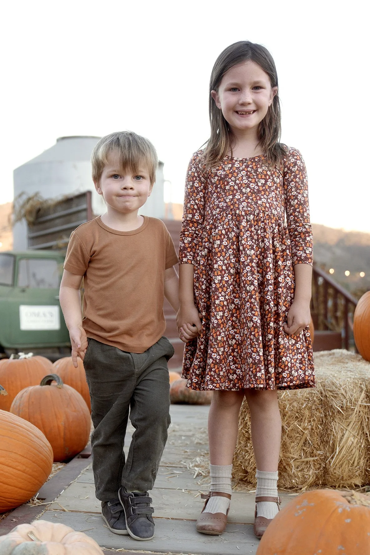 Two children, a girl and a boy, holding hands at a pumpkin patch with pumpkins and hay bales, during fall, with a farm truck in the background.