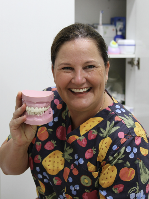 Smiling woman in colorful floral scrubs holding a dental model with teeth in a medical office or clinic.