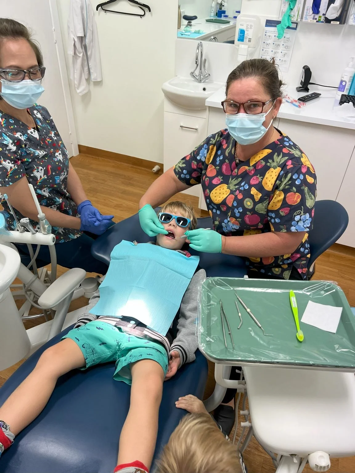 A young boy lying on a dental chair at the dentist's office, wearing sunglasses, receiving dental care from two female dental professionals. Both professionals are wearing masks, gloves, and colorful scrubs; one is standing at the head of the chair, and the other is adjusting the boy's mouth.