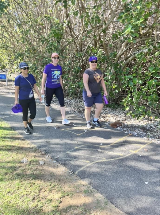 Three women walking on a paved trail surrounded by bushes, with chalk drawings on the ground and purple bags in hand.