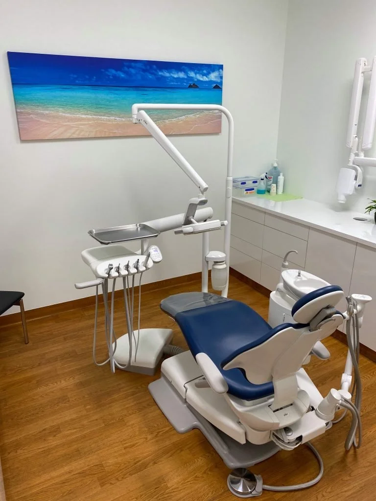 A modern dental examination room features a dental chair with attached equipment, a beige wall with a scenic beach poster, and a white cabinet with various dental supplies.
