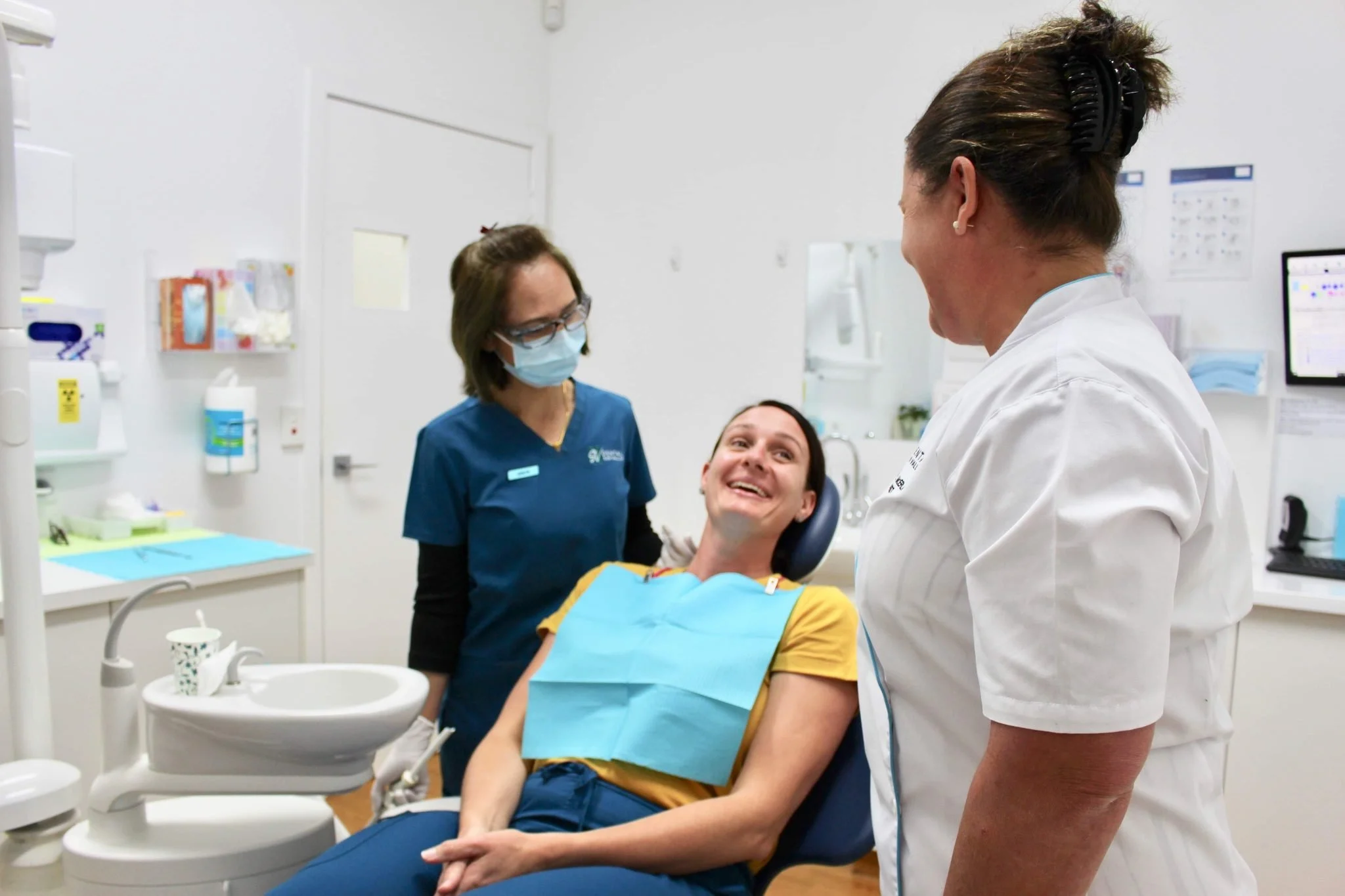 A woman in yellow shirt sitting in a dental chair smiling at a dentist and hygienist in a dental office.