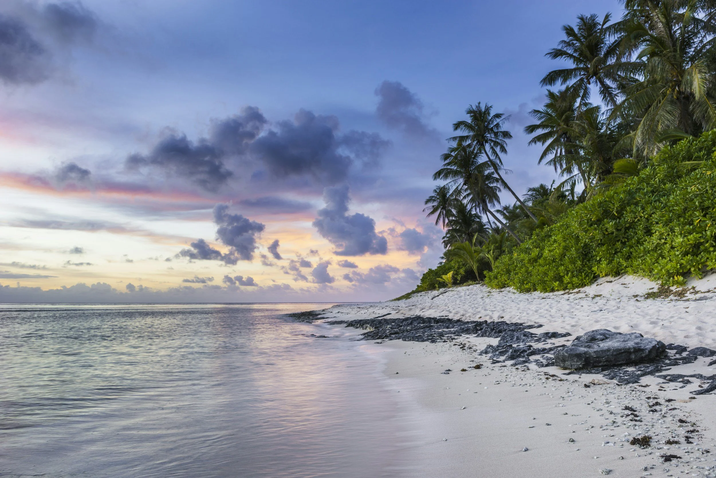 Sunset over a tropical beach with palm trees, green foliage, white sand, and rocks, with cloudy sky and calm water.